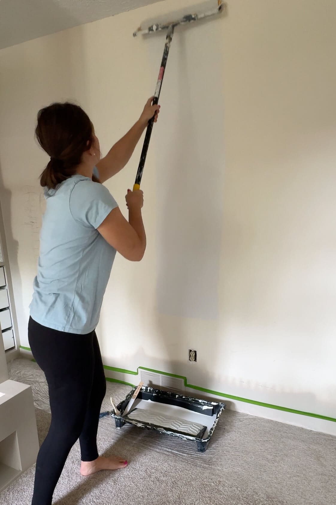 Woman using a paint roller with an extension pole to apply white primer to a beige wall.