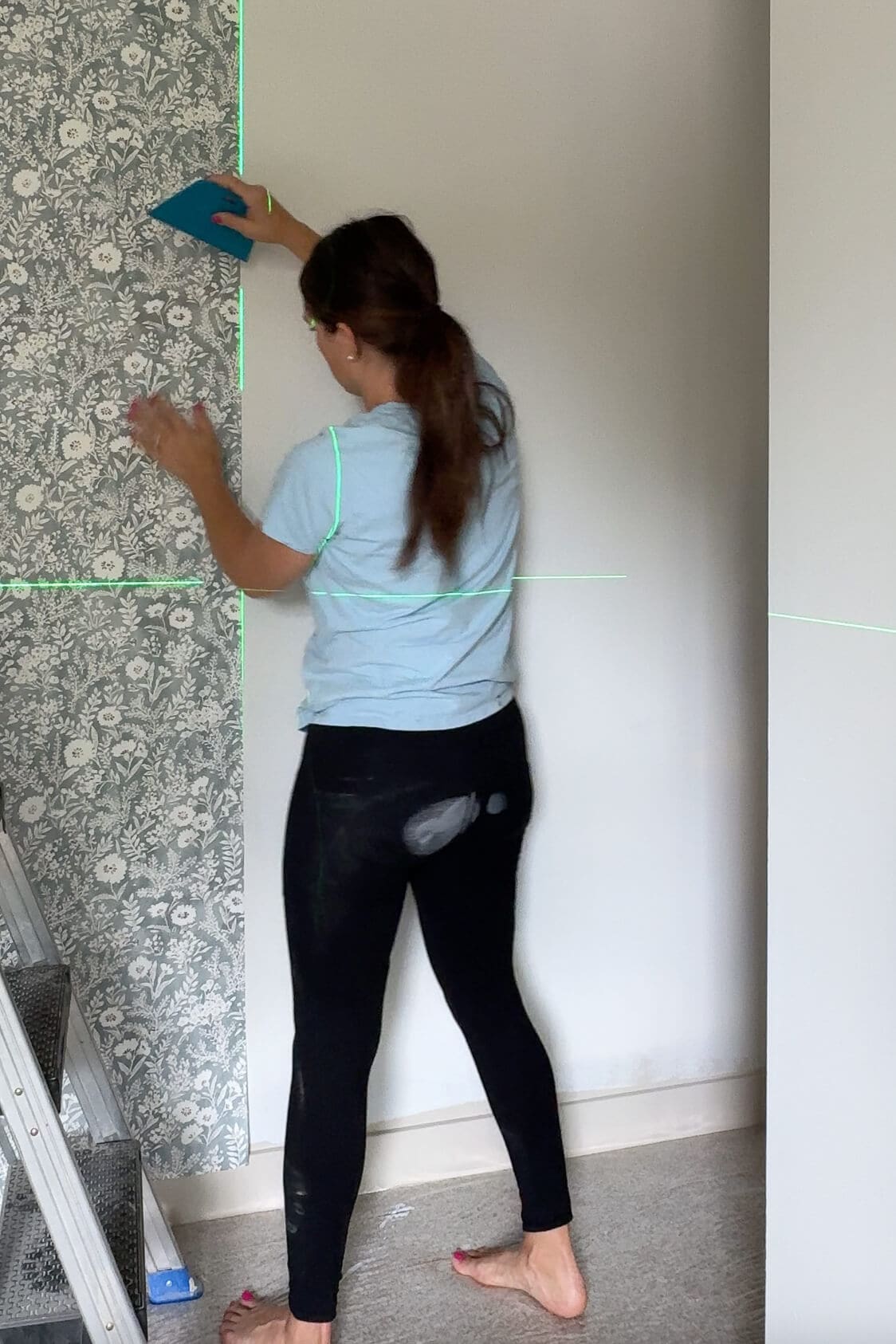 Woman smoothing a panel of wallpaper onto the wall with a plastic smoothing tool, aligning edges with a laser guide.