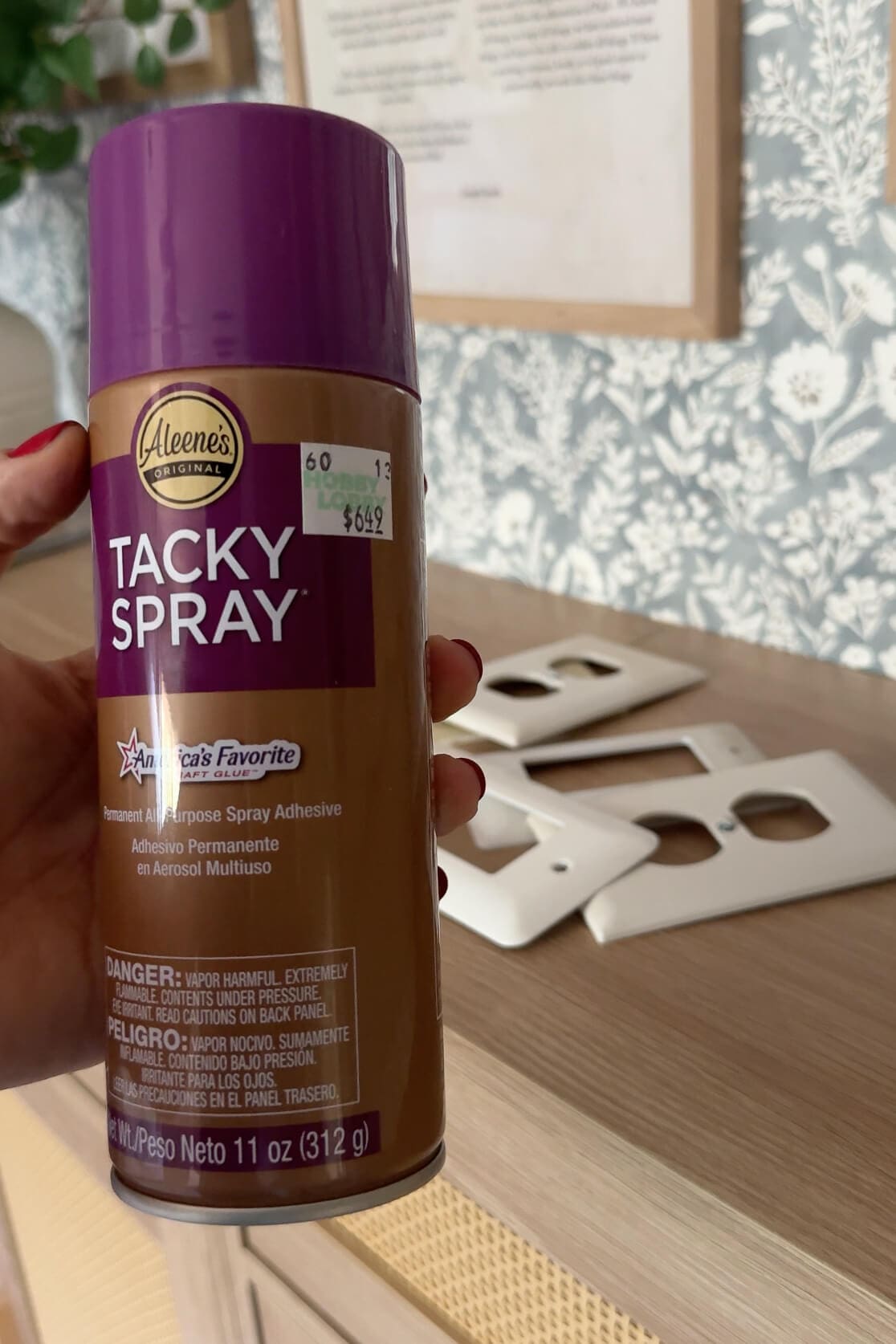 Aleene’s Tacky Spray adhesive held in front of several wall switch plates, used in a DIY project.