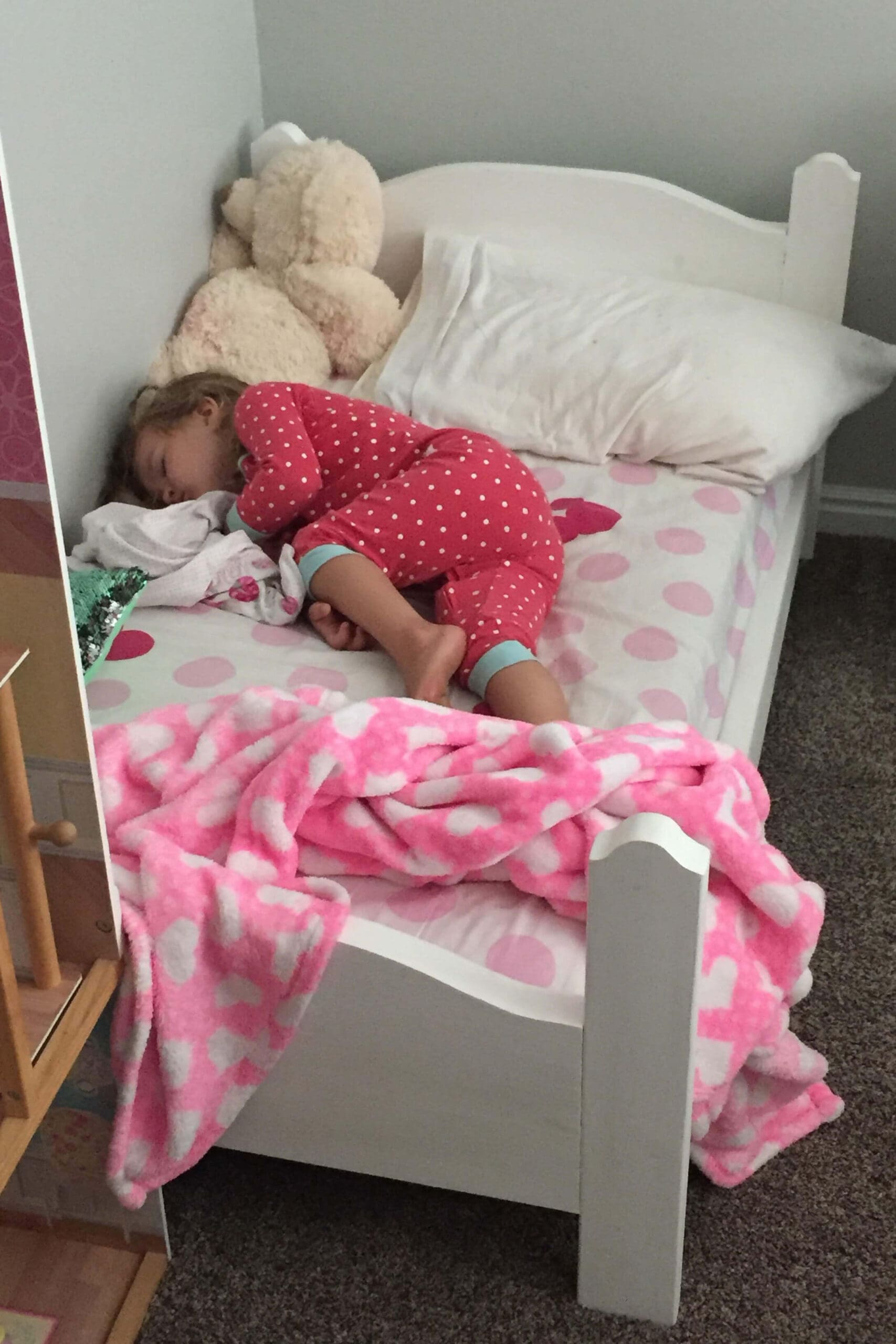 Toddler peacefully asleep in a handmade wooden bed with white paint and pink polka dot bedding, cozy under a soft pink blanket.