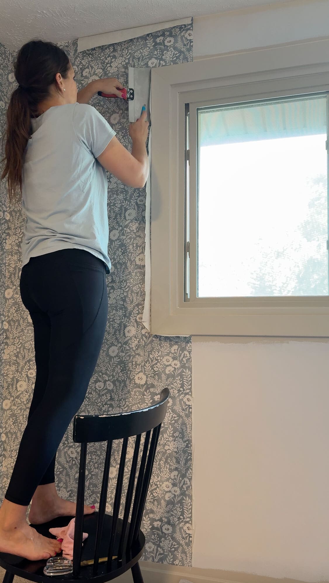 Standing on a chair, woman trims wallpaper along the top corner of a window using a smoothing tool and razor blade.