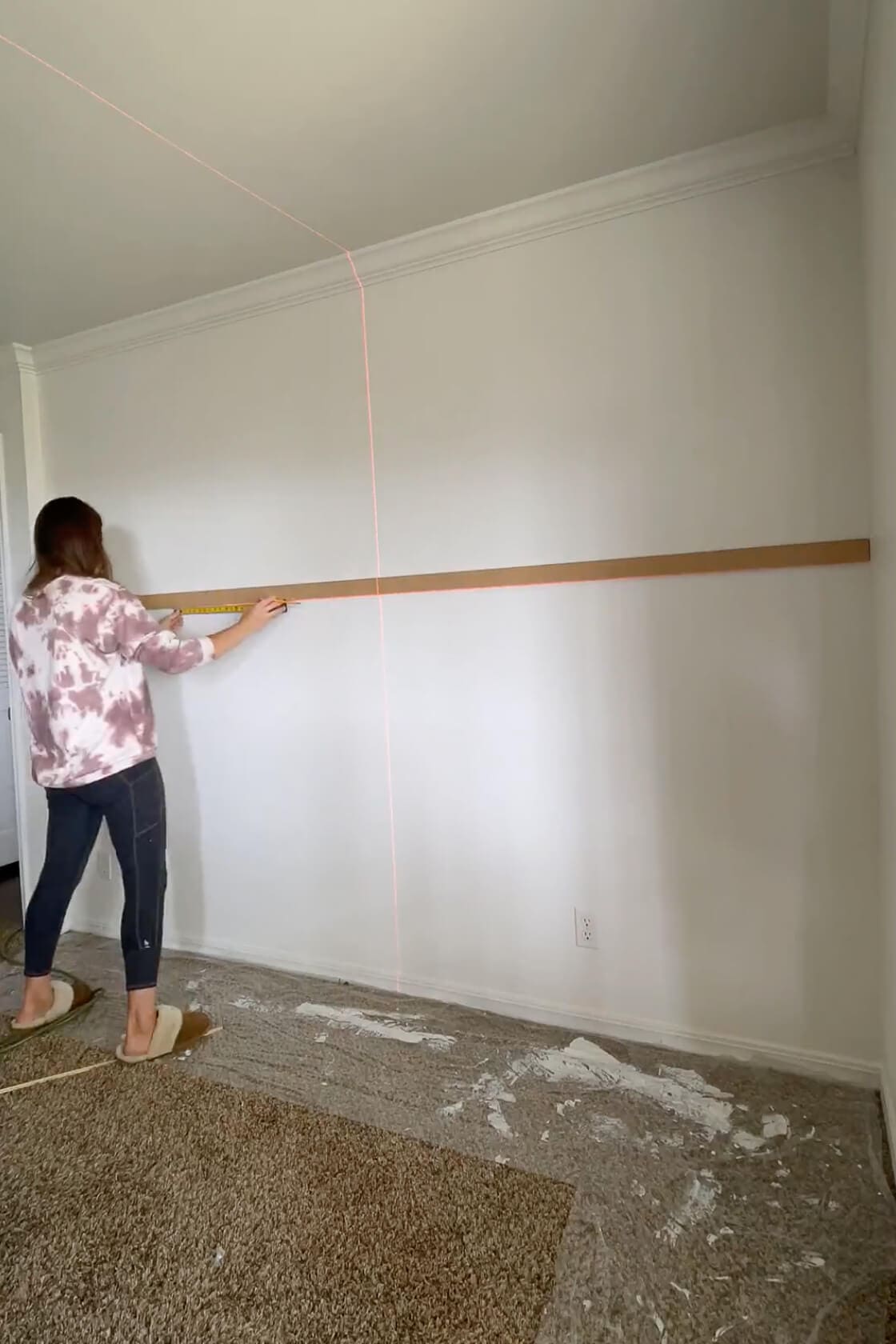 Woman measuring and marking a horizontal trim piece across a painted wall using a laser level to prep for more trim.