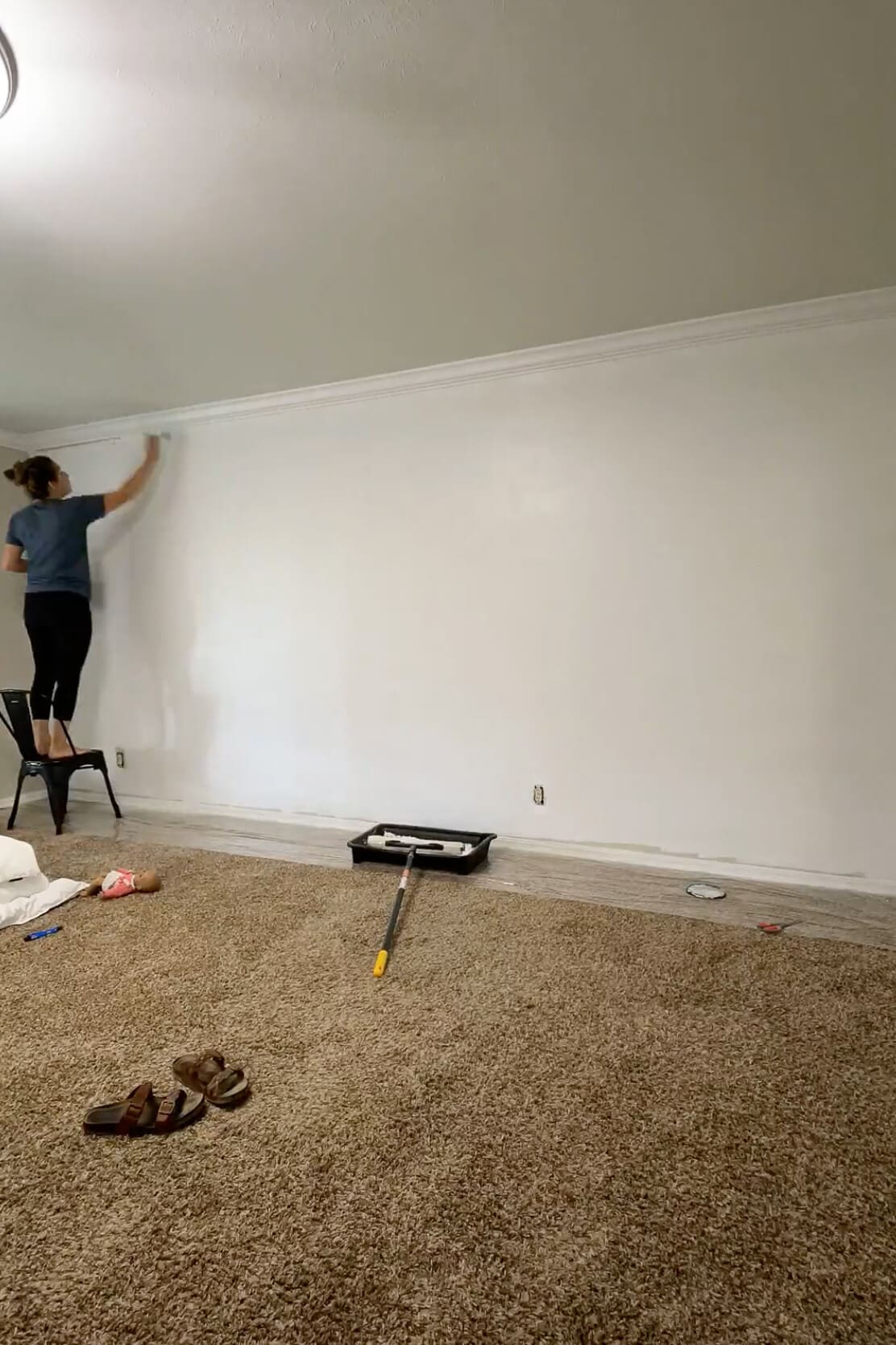 Woman using a roller to paint a wall white while standing on a chair