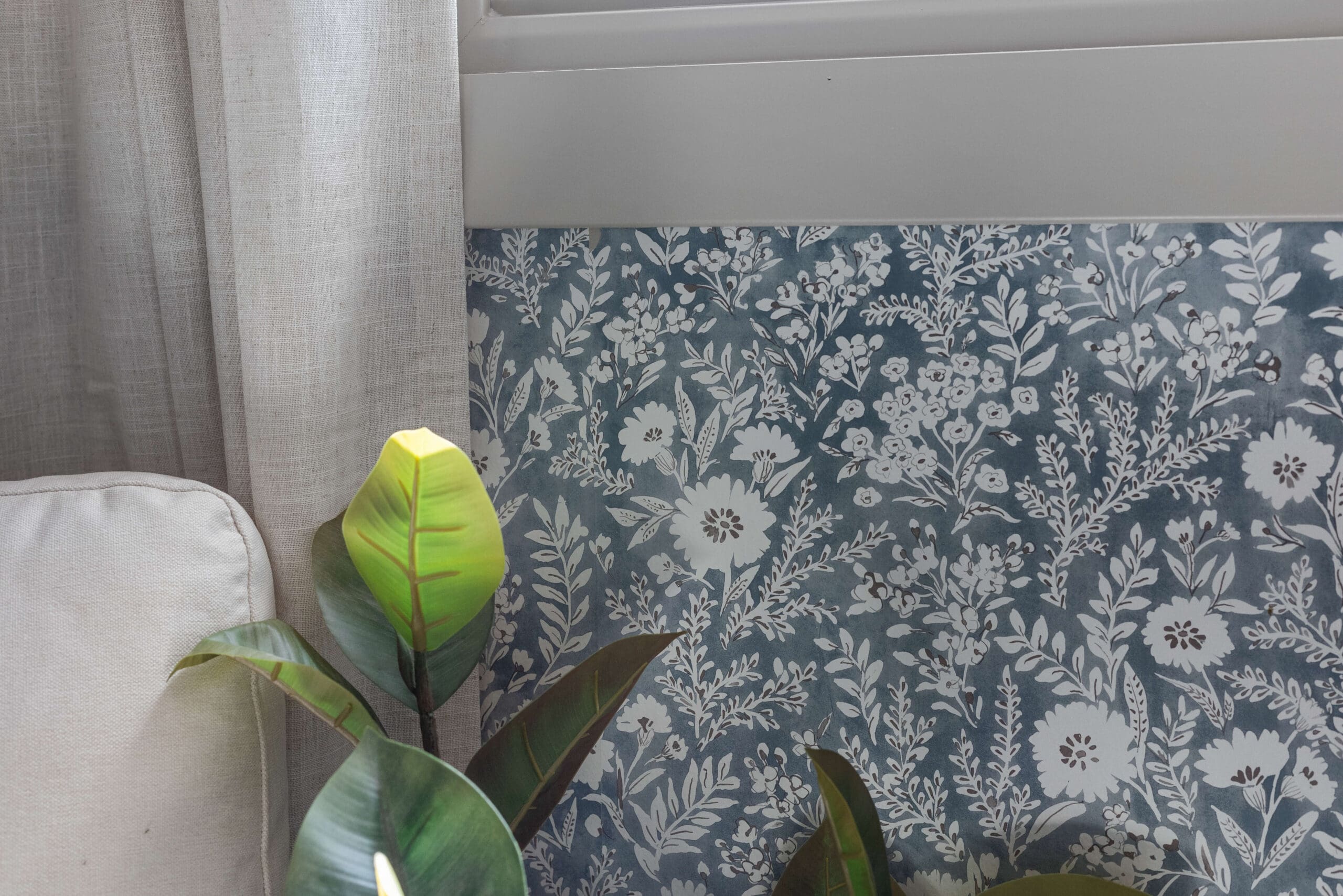 Detailed view of floral wallpaper beneath a window, next to linen curtains and a potted rubber plant.