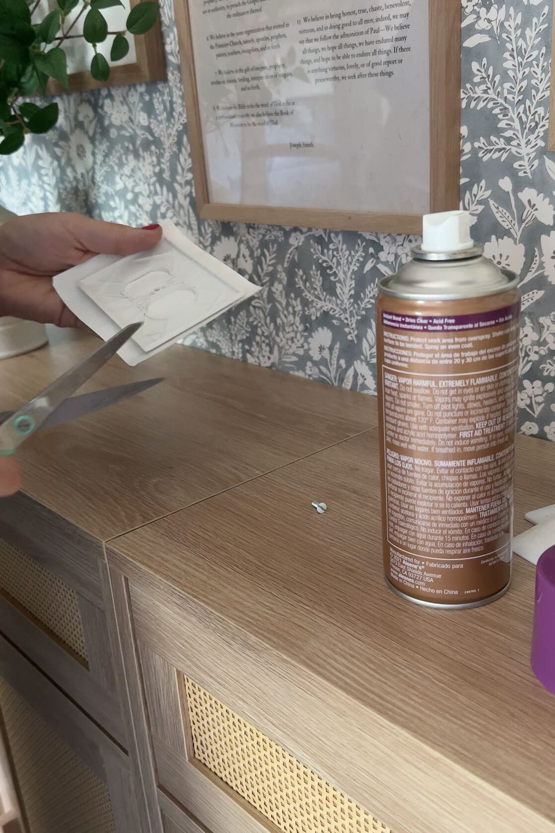 Trimming the edges of wallpaper wrapped around an outlet cover, with a can of spray adhesive on the work surface nearby.