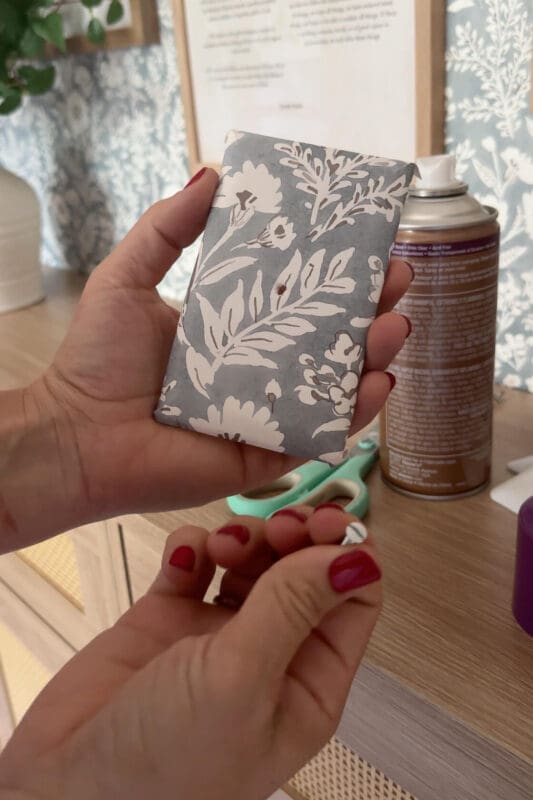 A finished wallpaper-covered switch plate held in hand, showing how the floral pattern wraps neatly around the edges for a polished finish.