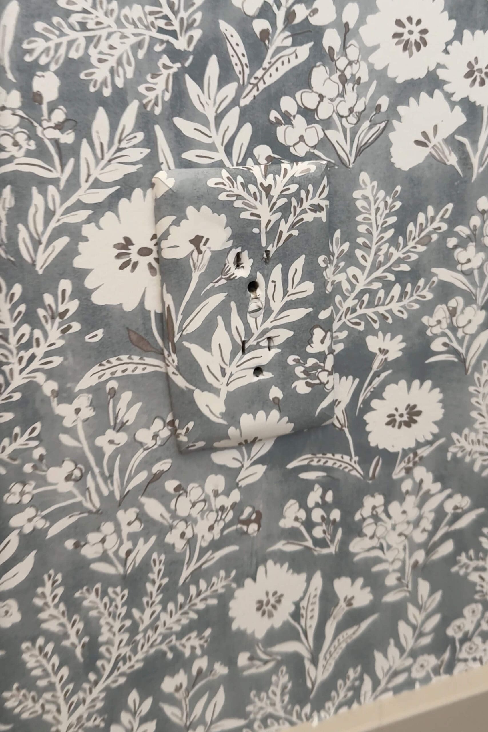 Close-up of a wallpapered outlet cover blending almost seamlessly into a floral patterned wall, with screw holes lined up for installation.