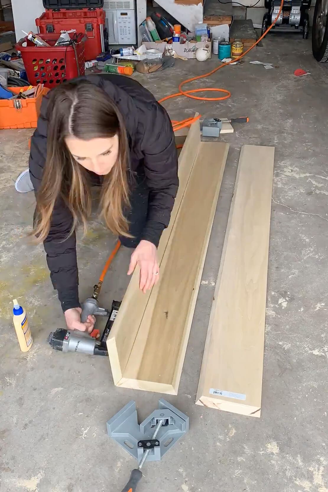 Woman building a DIY mantel by assembling wood boards with wood glue and a nail gun inside a garage workshop.