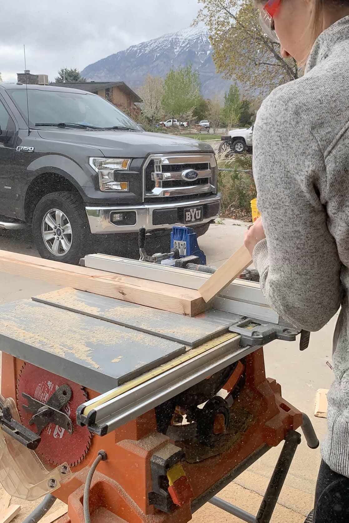 Cutting an angled wood leg on a table saw during a DIY coffee table build, with safety glasses on and a mountain view in the background.
