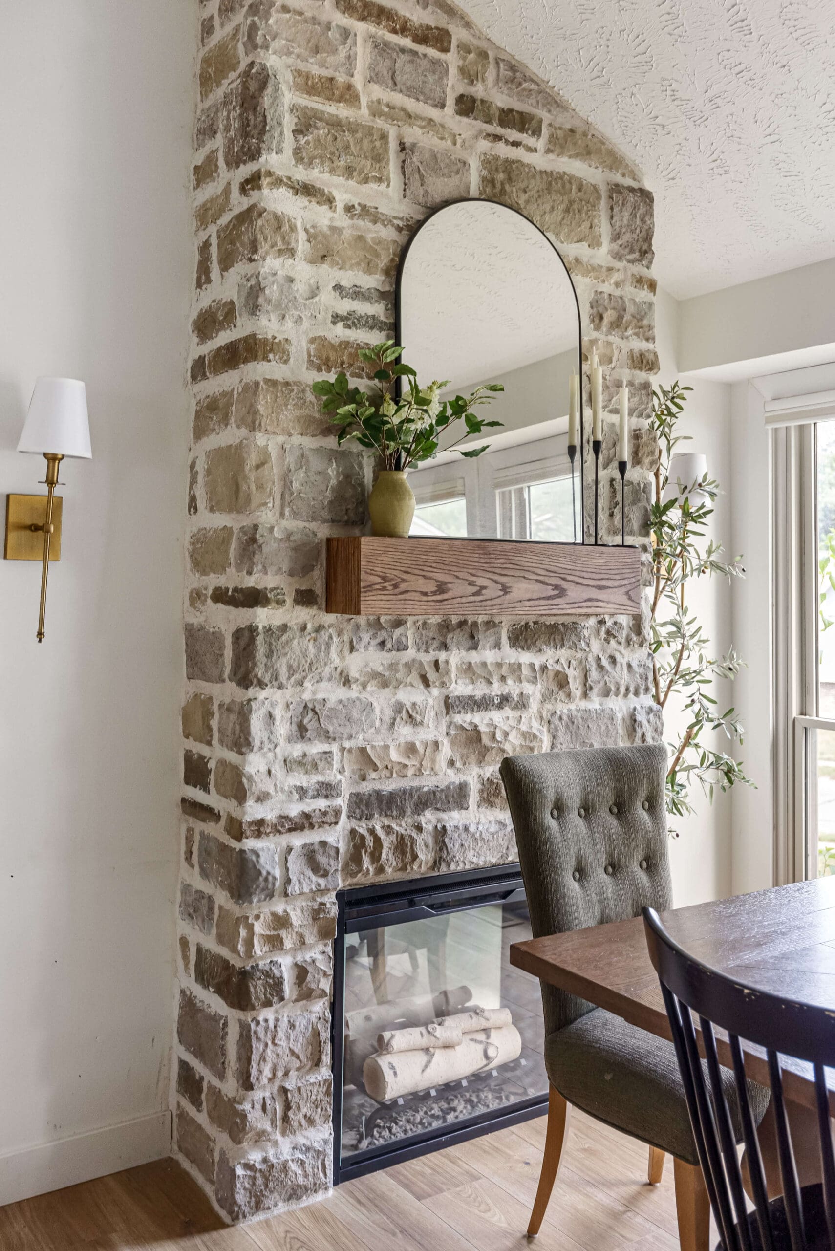 Floor-to-ceiling stone fireplace in a dining room, styled with a wood mantel, arched mirror, greenery, and modern candlesticks beside a dining table.