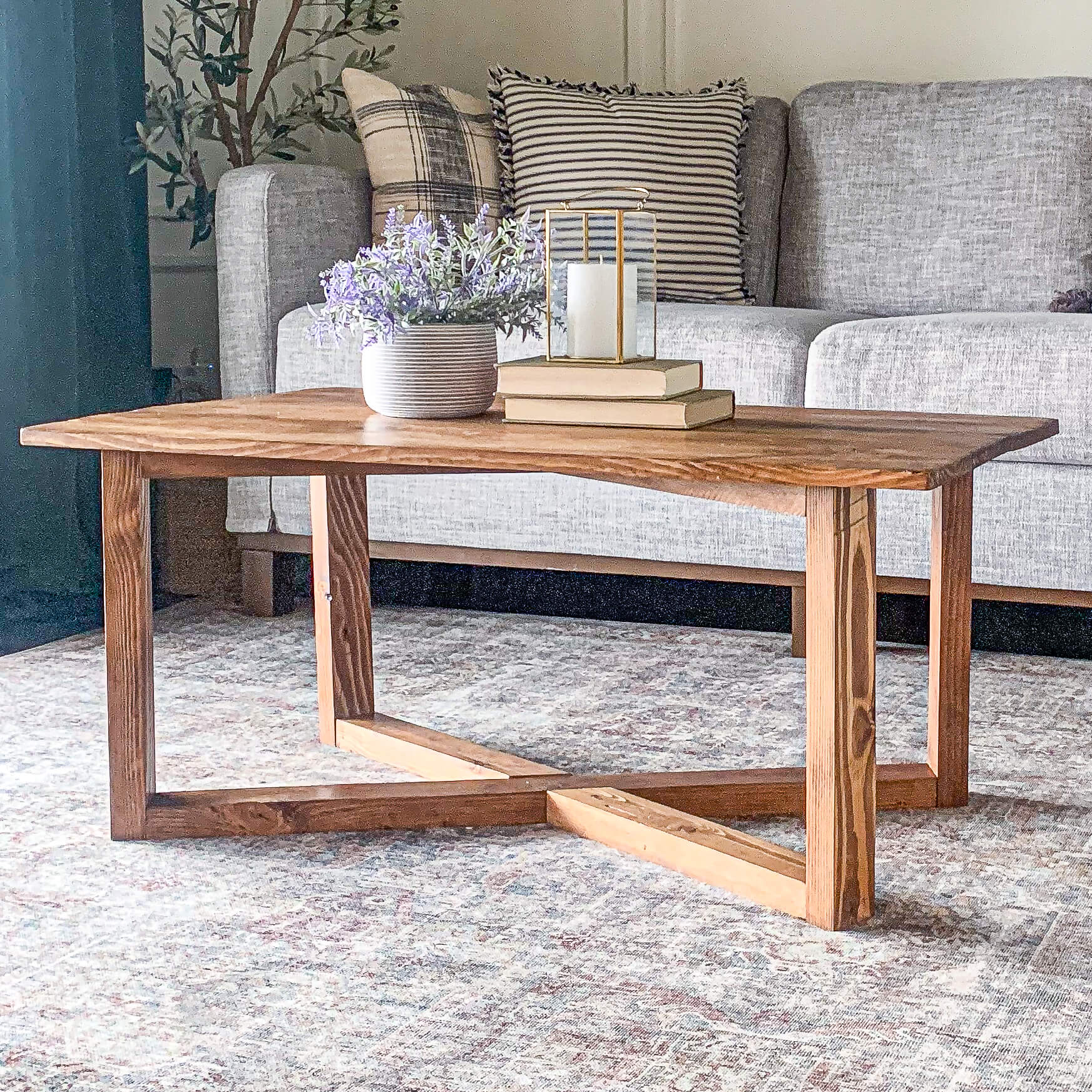 Handmade wooden coffee table with a modern geometric base, styled in a cozy living room with a gray couch, striped pillows, and a vintage-style area rug.