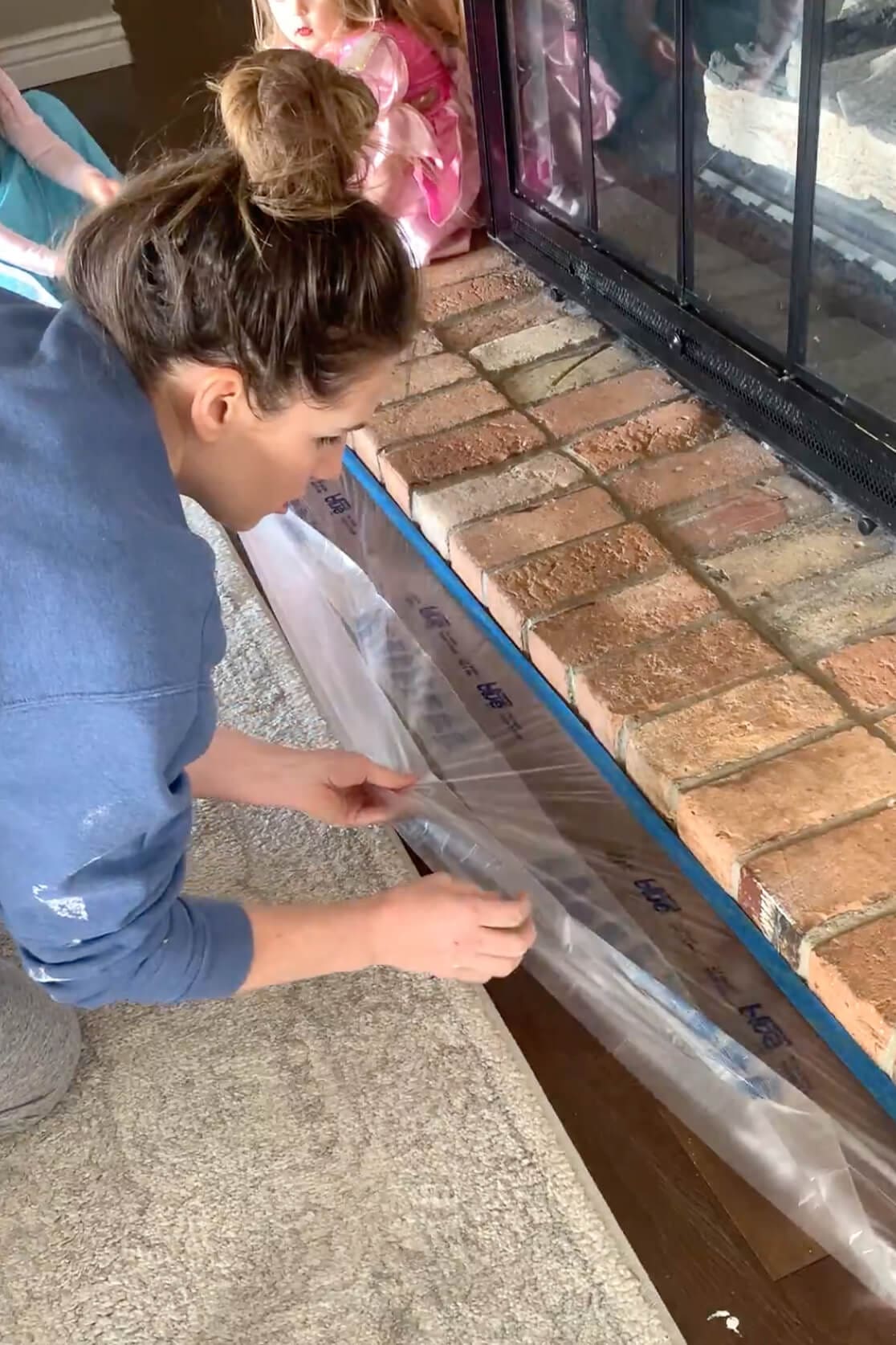 DIY fireplace update underway with masking materials applied to protect the floor and firebox; brick surface partially covered and prepped.