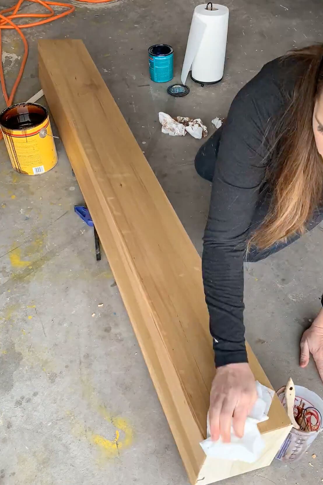 Close-up of a DIY wood mantel being stained by hand in a garage, with cans of stain and supplies visible nearby.