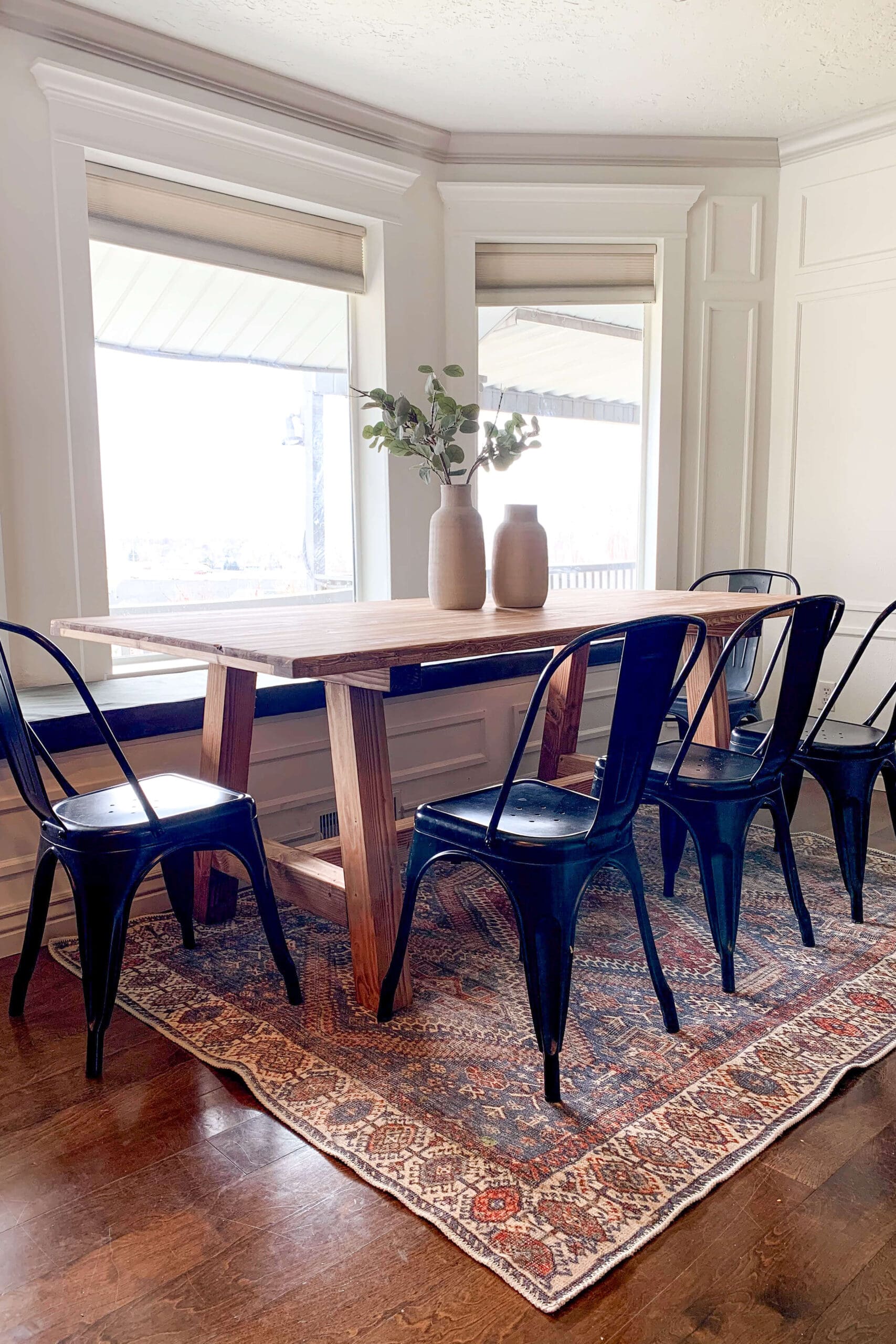 DIY wooden dining table with a trestle base, styled with black metal chairs, wood vases, and a vintage rug in a bright bay window nook.