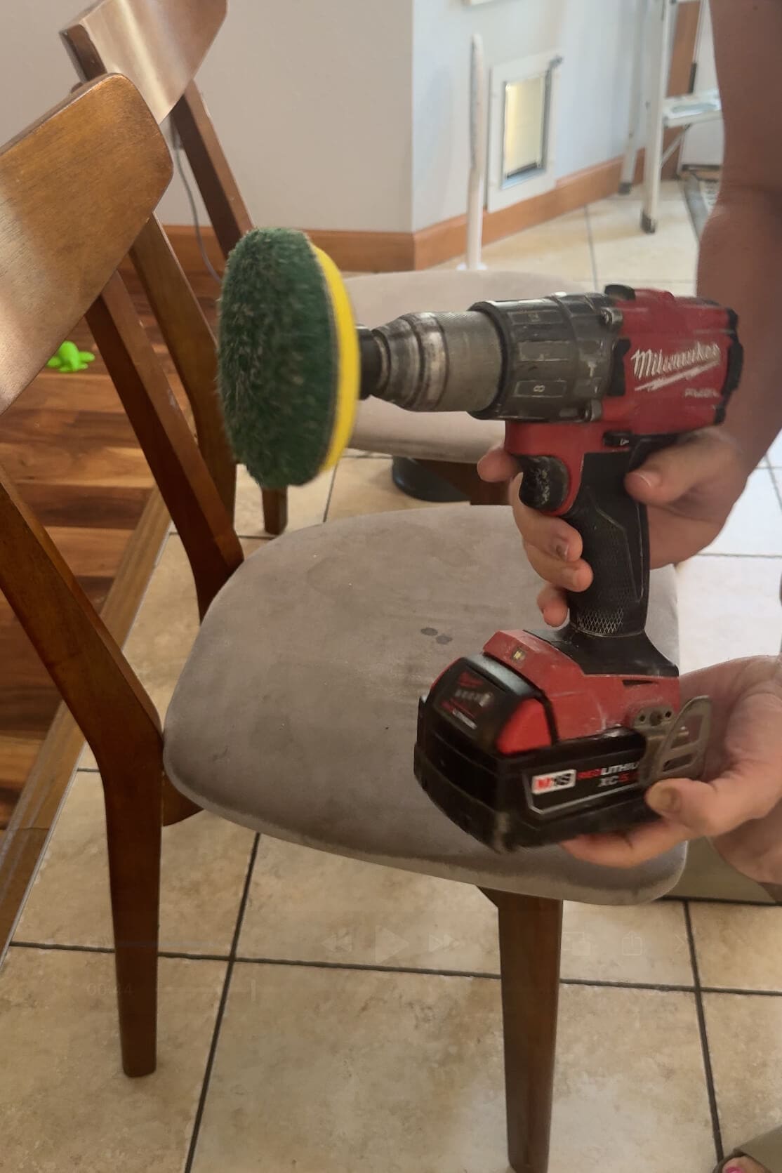 Holding a power drill with a green scrub brush attachment before cleaning a fabric dining chair.