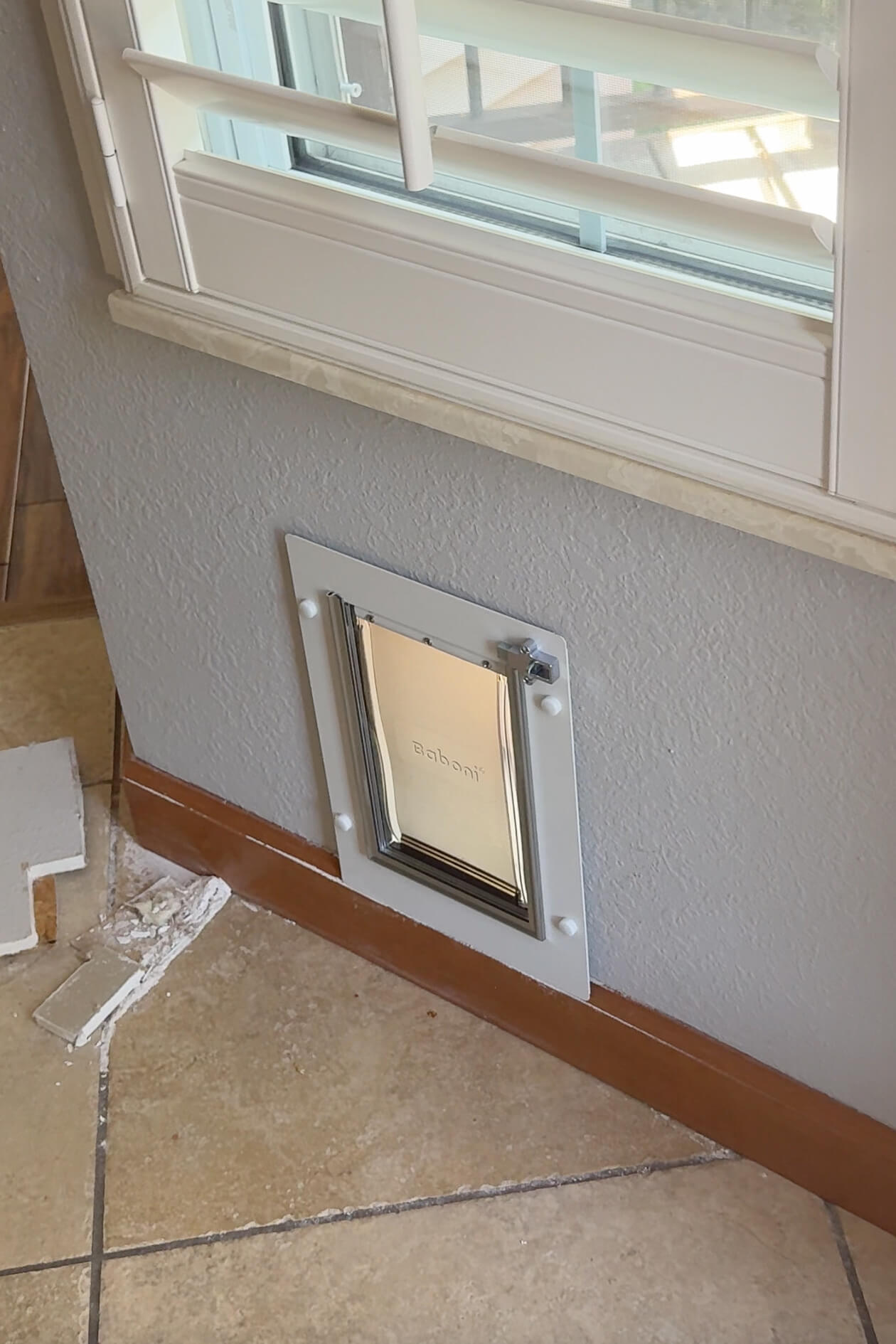 Completed dog door installation on an interior wall beneath a window with plantation shutters and tile flooring.