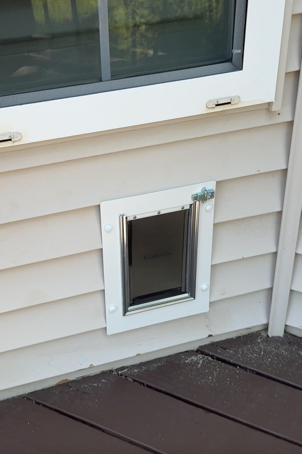 Close-up view of a white-framed dog door installed on tan vinyl siding beneath a window.