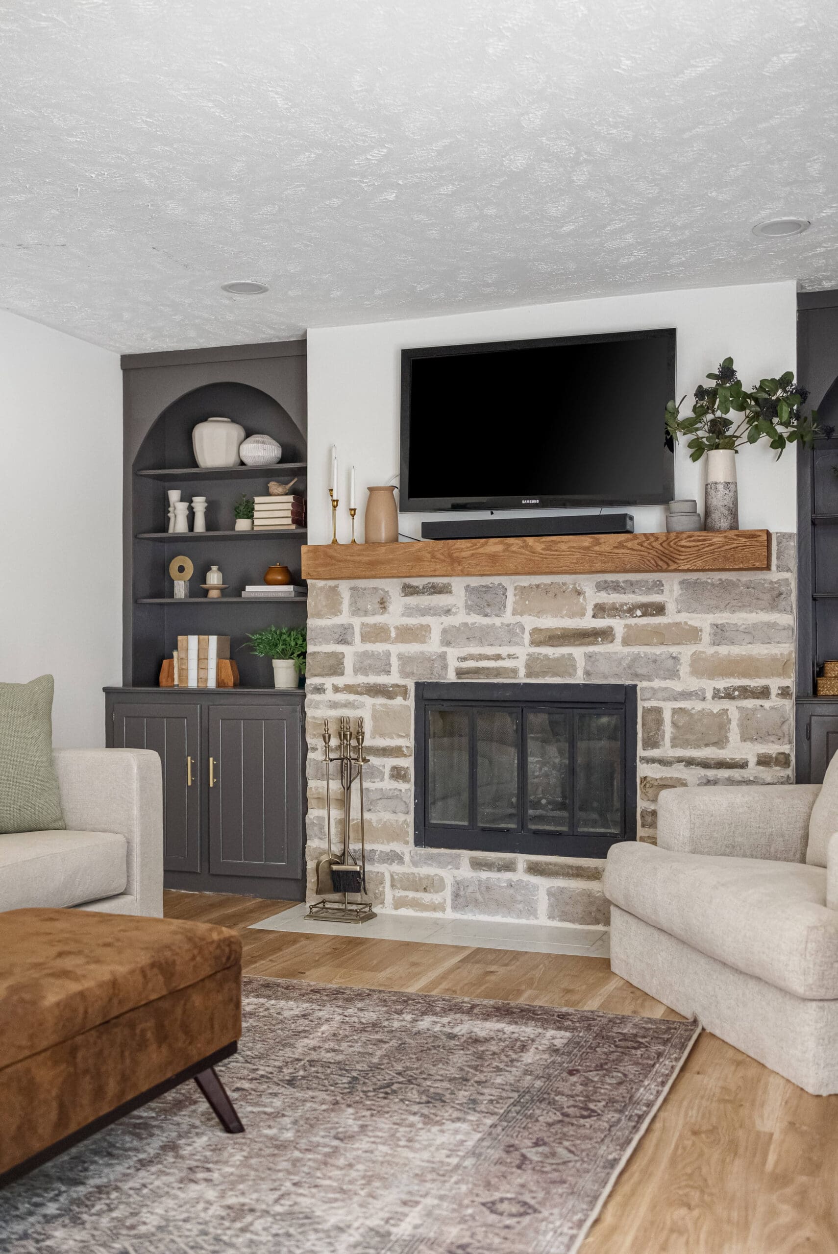 Stone fireplace with a wood mantel and mounted TV, flanked by dark built-in shelves in a cozy living room with neutral furniture and warm wood floors.
