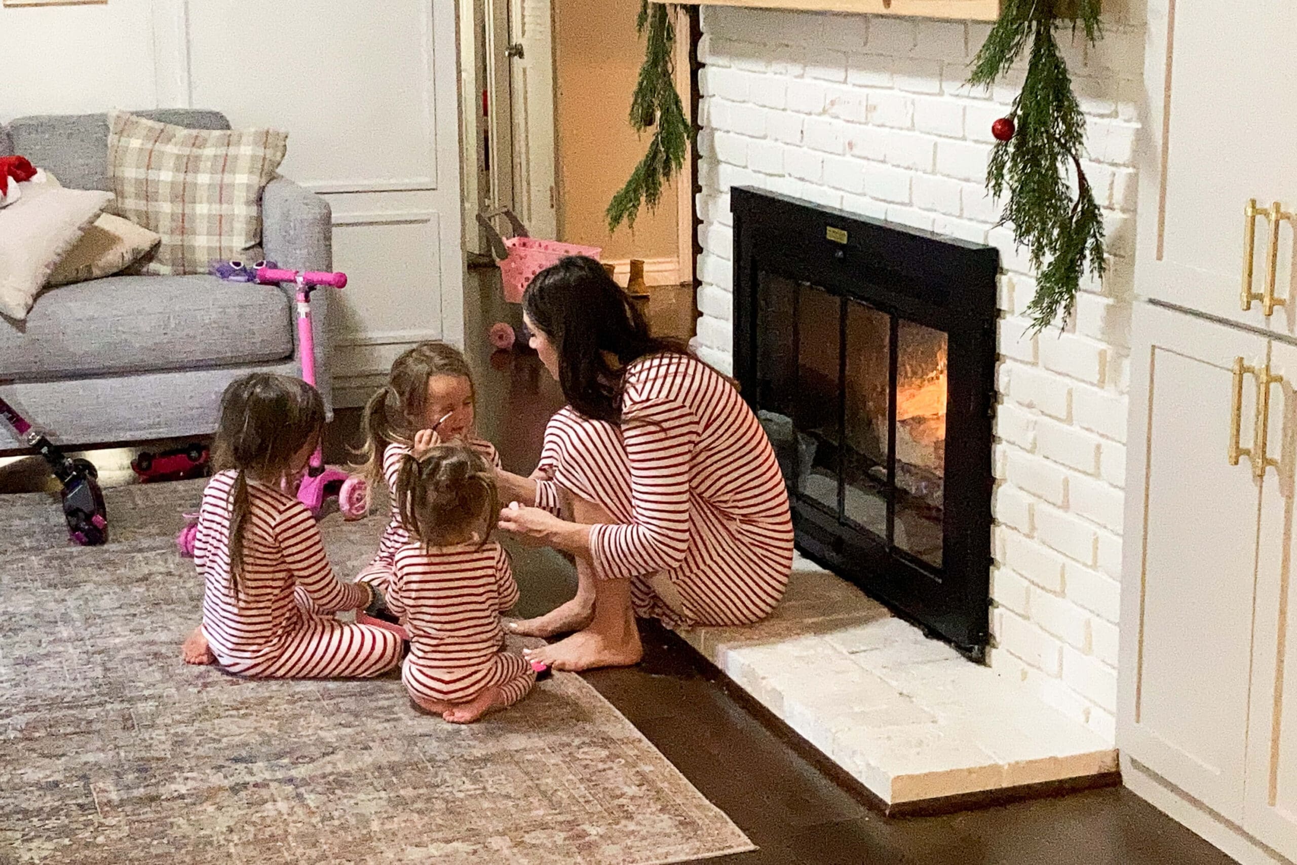 Mom and kids in matching striped pajamas gathered around a cozy fire beside a painted brick fireplace decorated with holiday greenery.