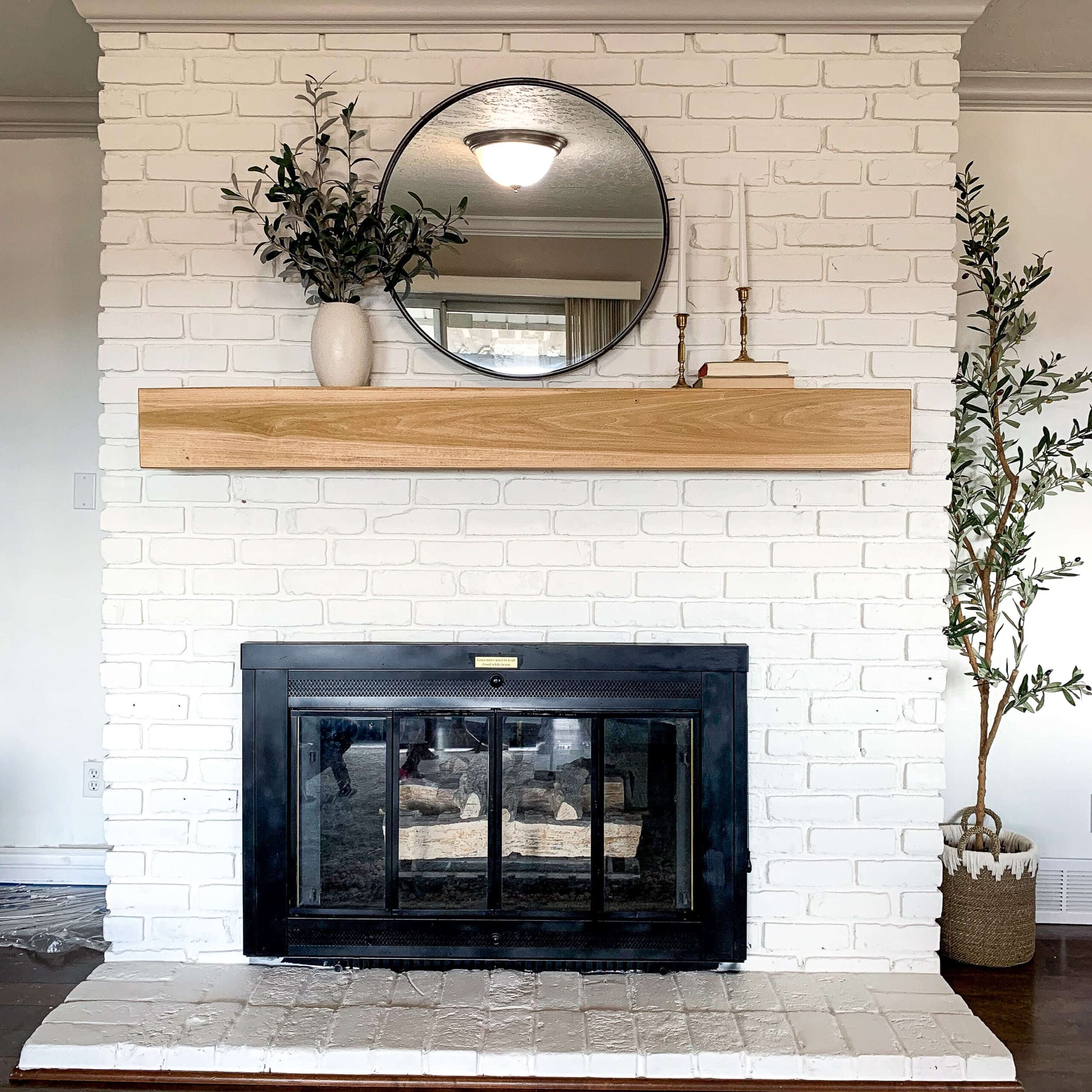 Painted brick fireplace in crisp white with a light wood mantel, round black-framed mirror, and minimalist decor including greenery and candlesticks.