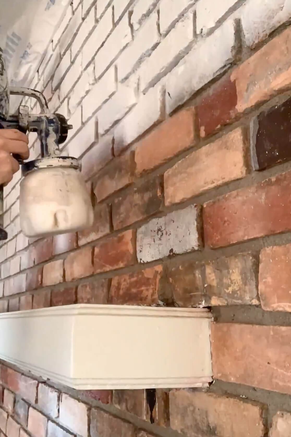 Close-up of a DIYer using a paint sprayer to coat red brick with white paint during a painted brick fireplace transformation.