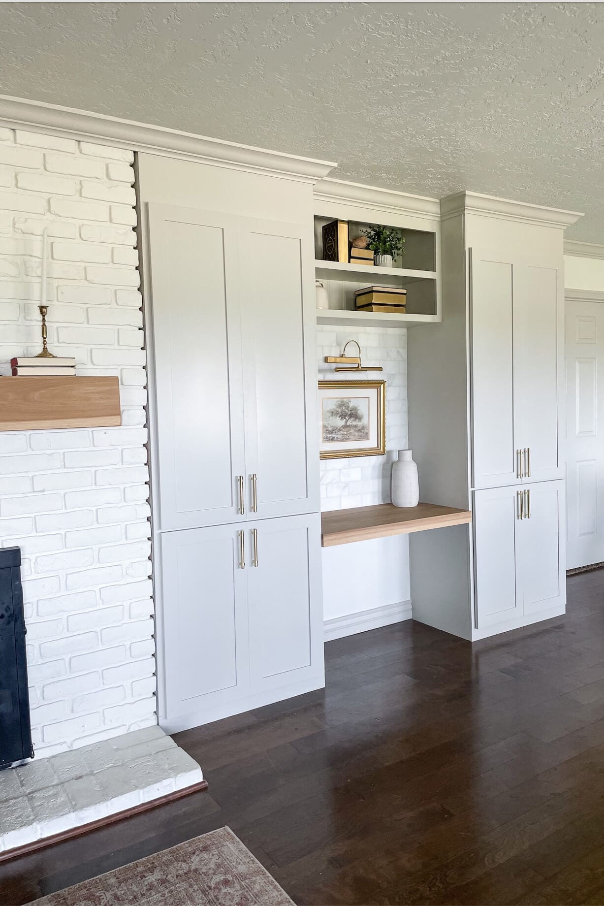 Freshly updated living room with white built-in cabinets and desk space next to a painted brick fireplace with a light wood mantel.