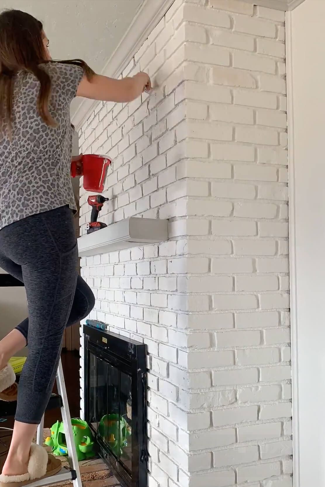 Woman on a ladder applying finishing touches to a painted brick fireplace using a brush, with white brickwork and a clean, updated look.