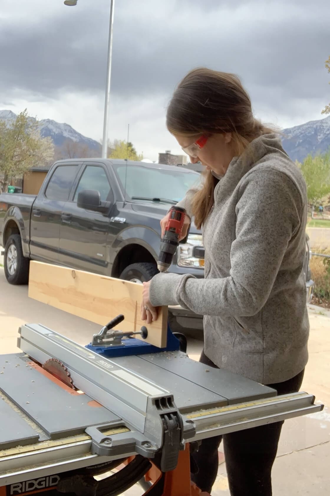 Using a pocket hole jig and drill to prep a wood board for joinery, with a table saw and truck in the background.