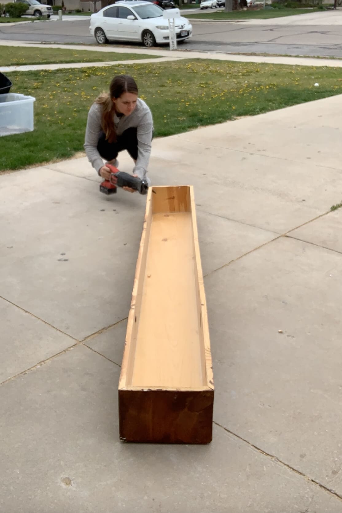 Woman using a reciprocating saw to dismantle an old wood fireplace mantel on a driveway, preparing to repurpose it for a DIY project.