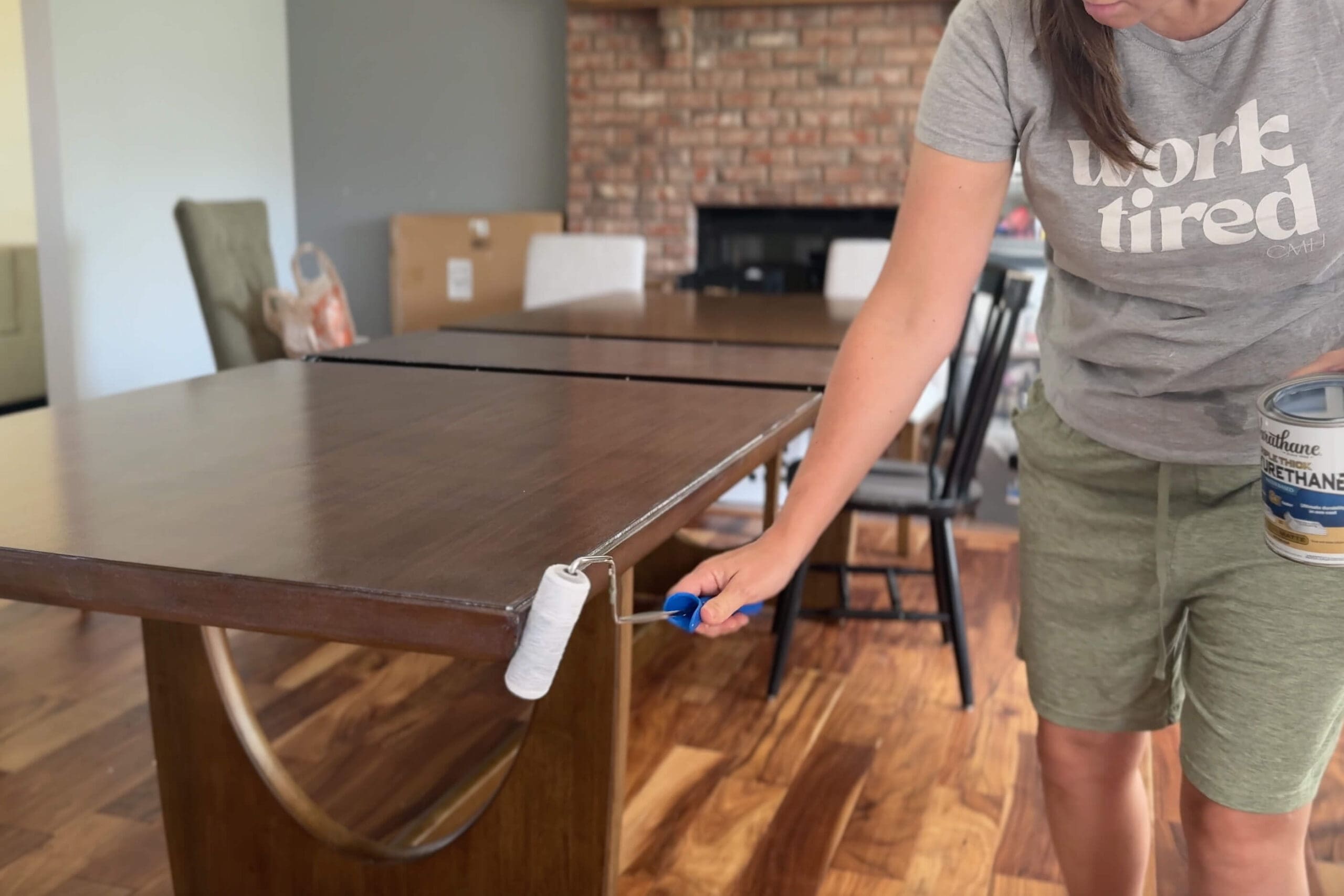 Applying polyurethane to the edge of a wood dining table using a foam roller, while holding a can of Varathane sealer in the other hand.