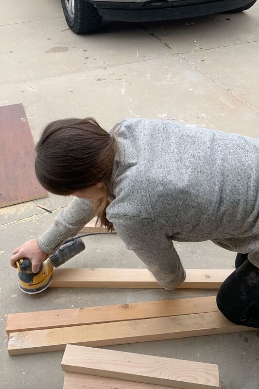 Sanding raw wood boards with an orbital sander to prep for staining and assembly during a DIY coffee table project.