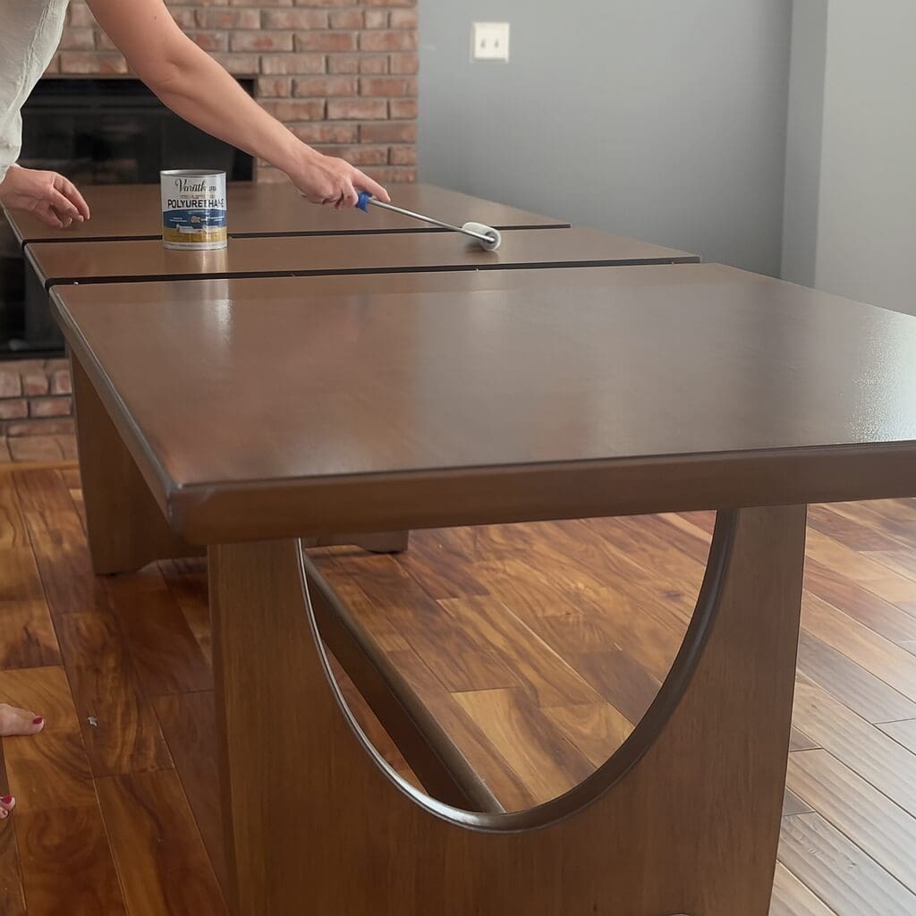 Woman applying polyurethane with a foam roller to seal a wood dining table, protecting the surface with a clear finish in a clean, modern dining room.