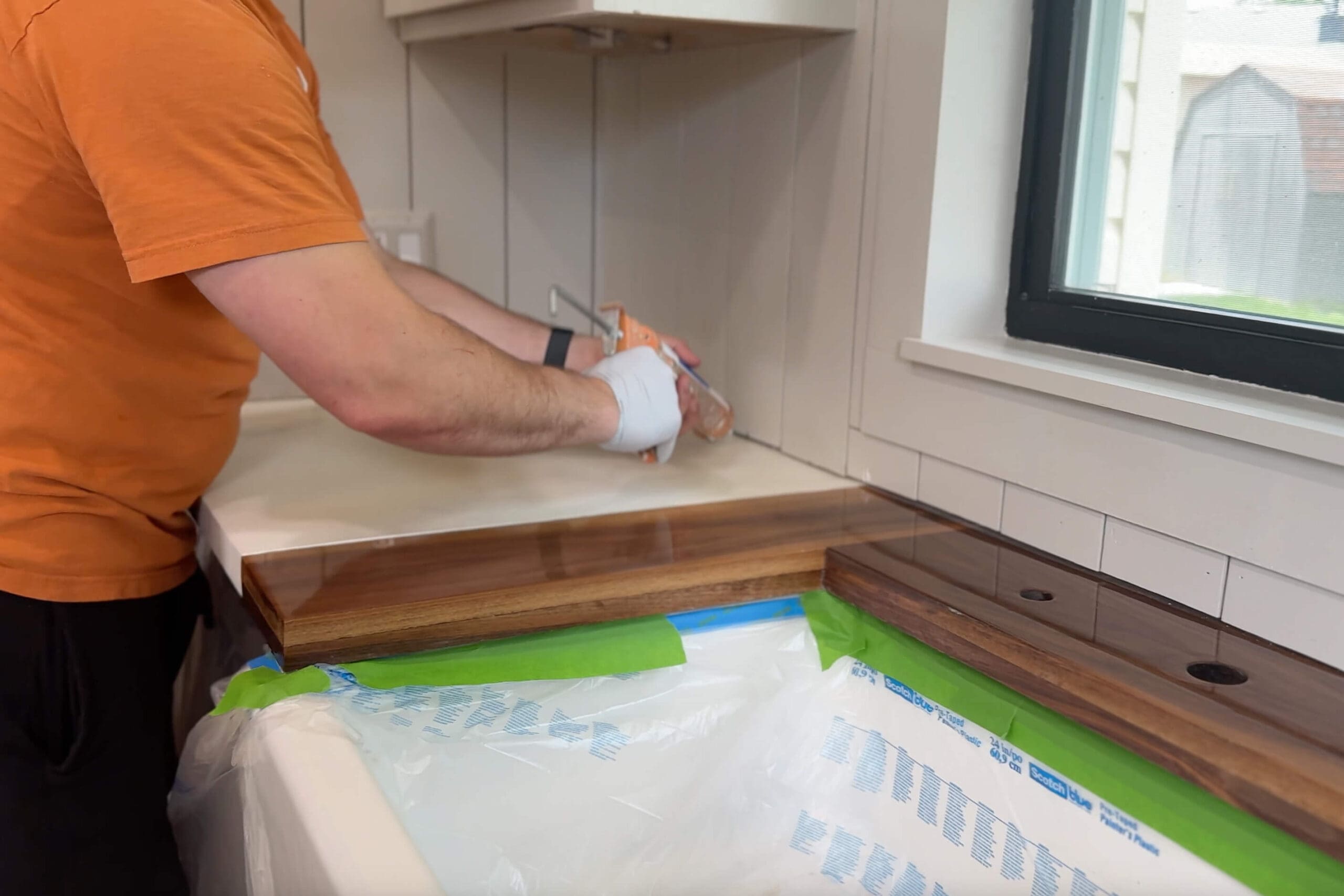 Sealing the edge of faux marble DIY counters with caulk next to a newly installed walnut trim piece around a farmhouse sink
