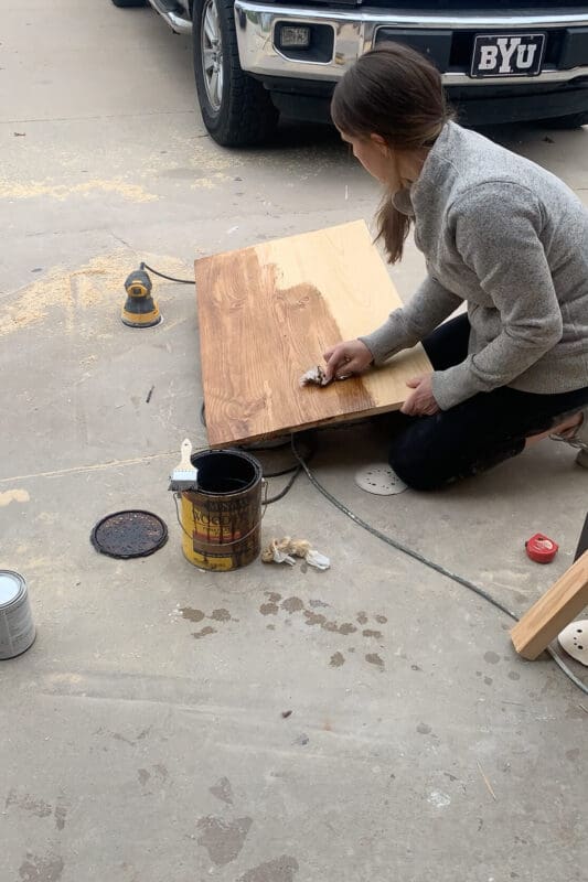 Applying wood stain to a DIY coffee tabletop made from joined boards, with an orbital sander, stain can, and painting supplies nearby on the driveway.