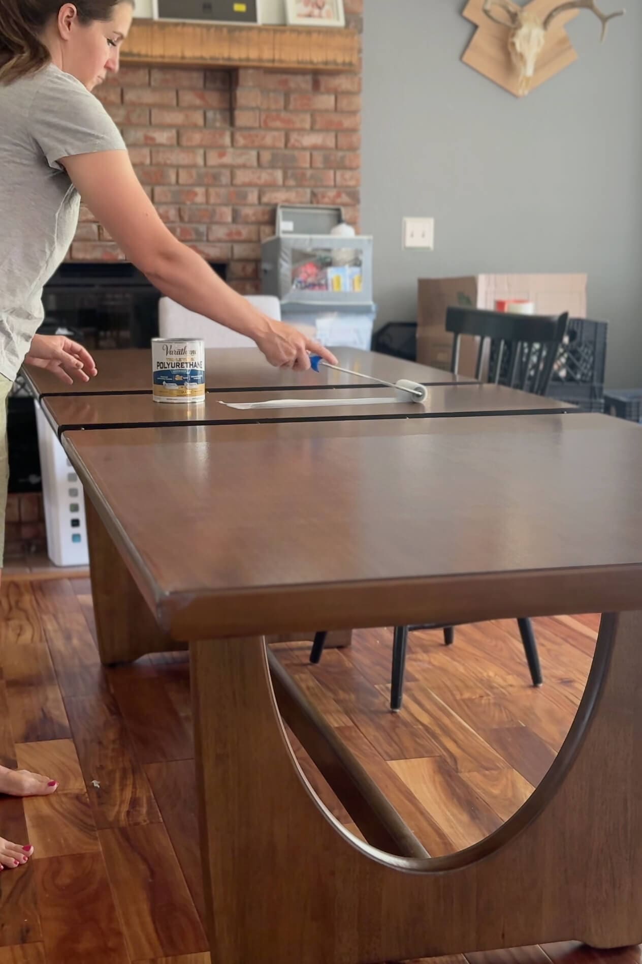 Woman applying polyurethane to a wood dining table with a foam roller, mid-process with the Varathane can placed on the tabletop.