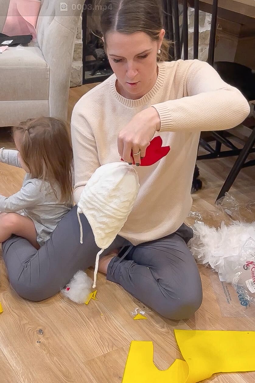 Mom attaching a red felt comb to a white knit hat while children cut batting and prepare other pieces for a chicken costume.