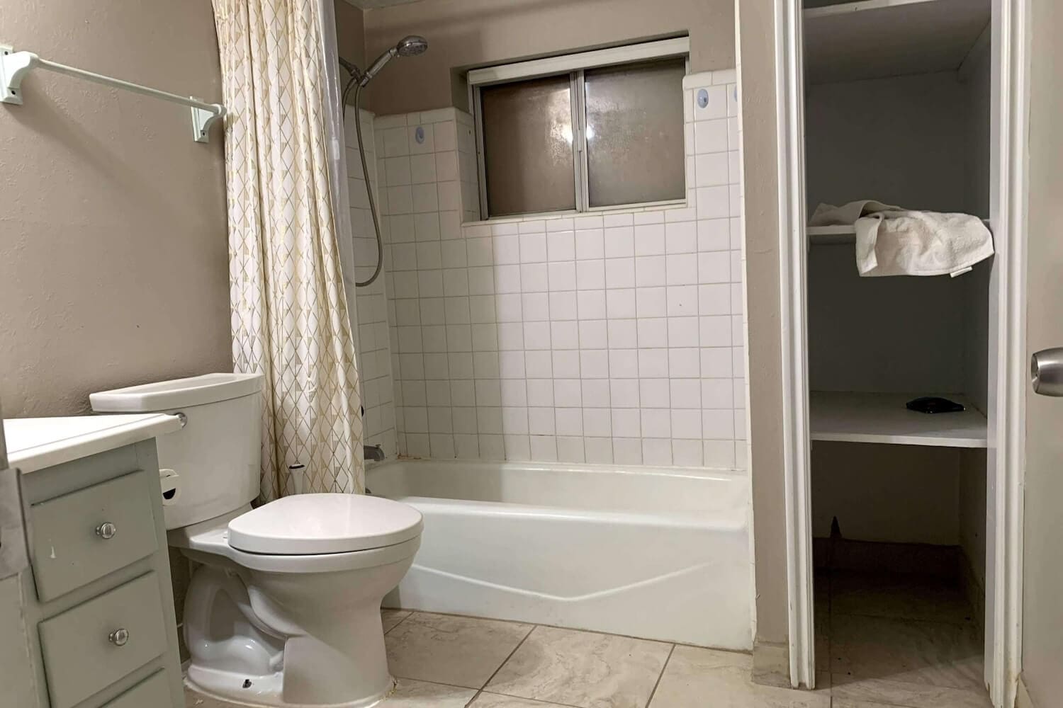 Basement bathroom before renovation with beige shower surround, outdated tile, and a small window inside the shower wall.