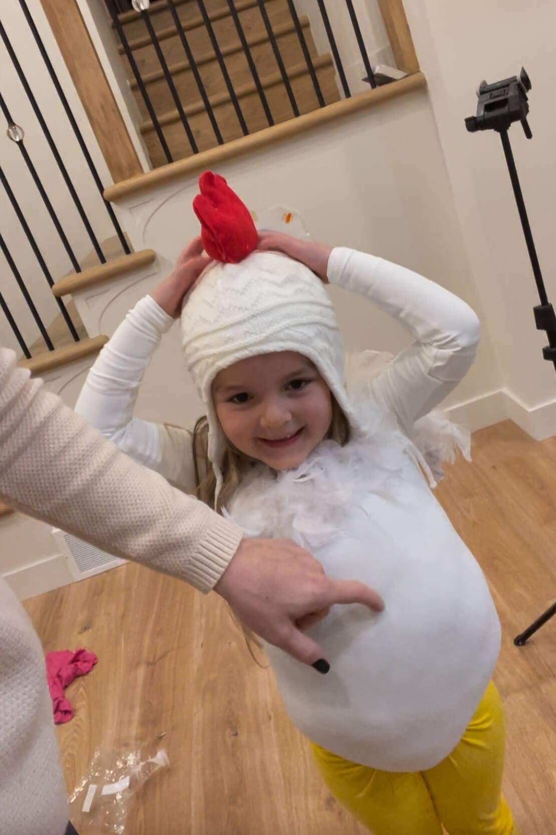 Smiling child wearing a white knit hat with a red felt comb and feathered shirt while trying on a chicken costume in progress.