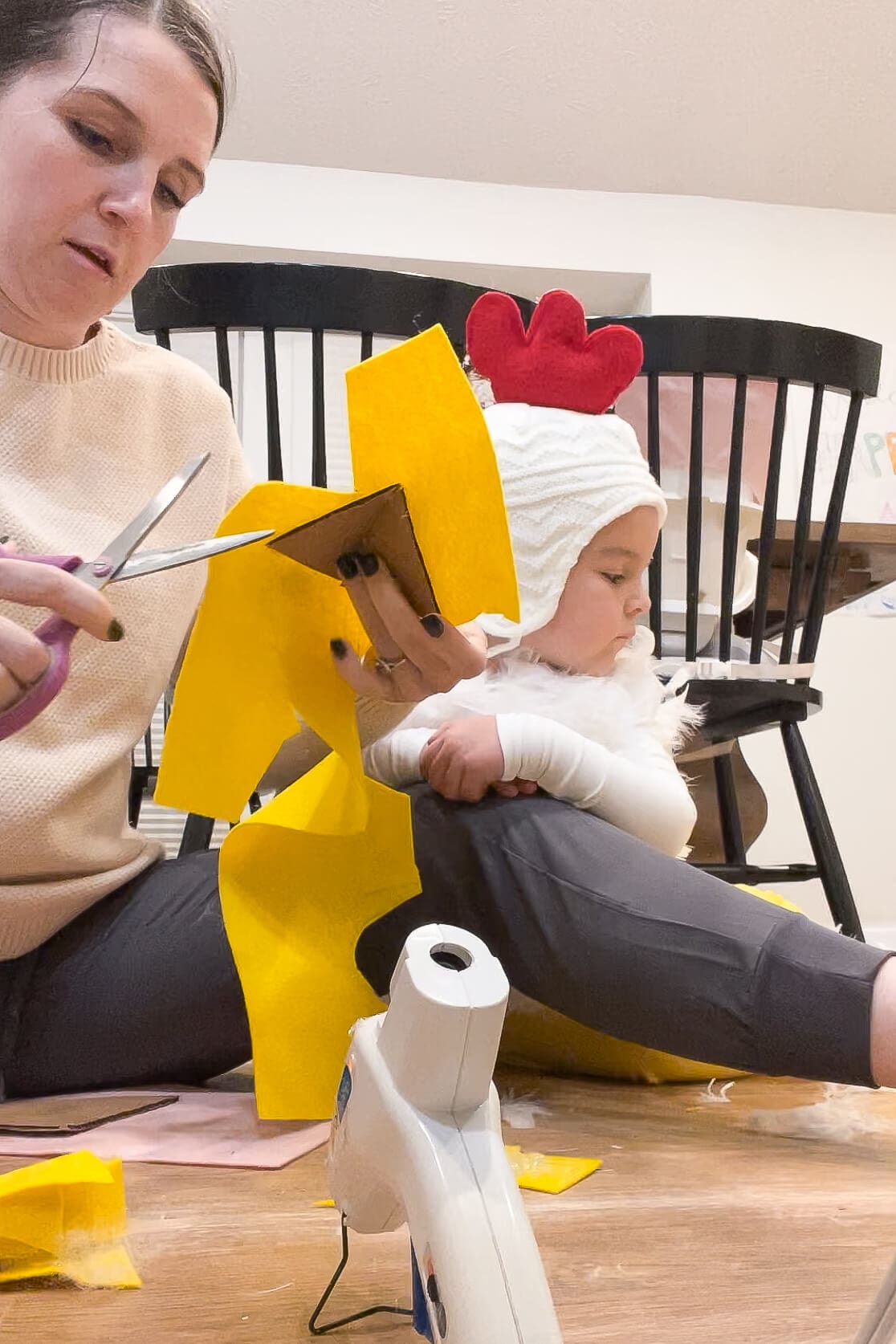 Mom cutting yellow felt to cover a cardboard triangle while a child dressed as a chicken sits nearby.