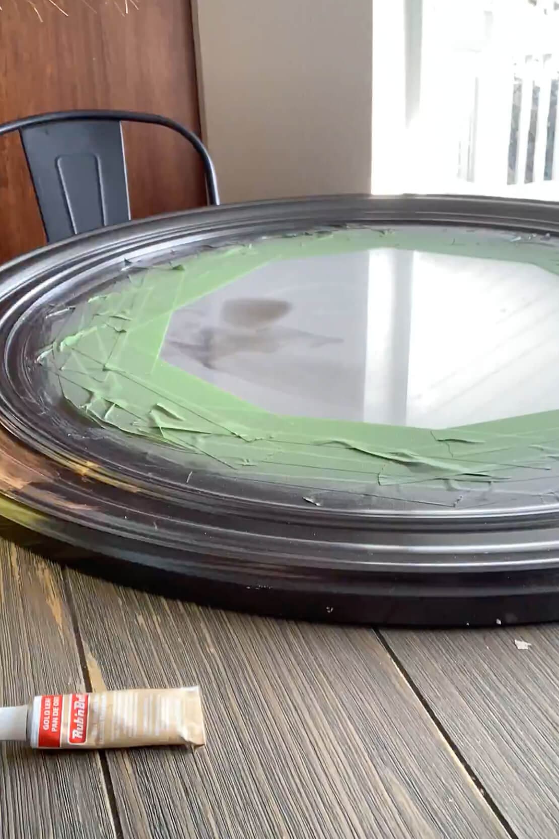 Round bathroom mirror lying flat on a table with glass taped off and a tube of Antique Gold Rub ’n Buff ready for a gold frame makeover.