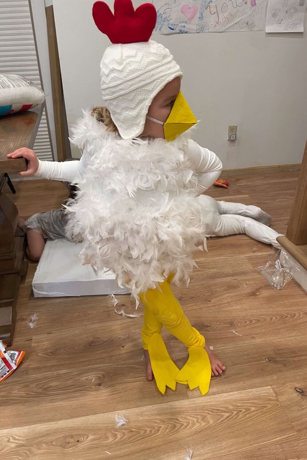 Side view of a child in a DIY chicken costume with white feathers, yellow leggings, knit hat, and felt beak
