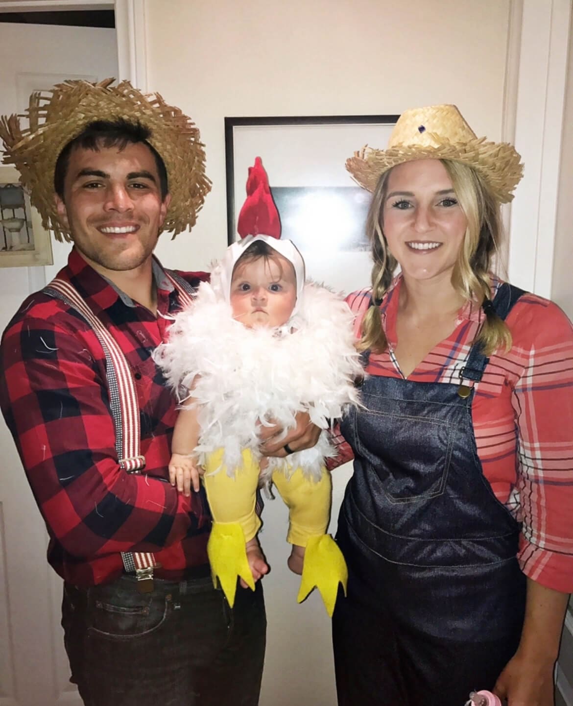 Family of three in matching themed family costumes with a barnyard theme — mom and dad dressed as farmers holding their baby in a no sew chicken costume, perfect for a country-inspired look.