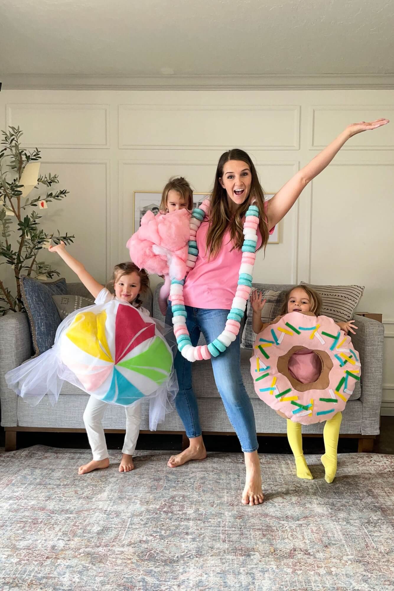 Mom and kids in matching candy costumes, featuring a giant candy necklace, cotton candy, lollipop, and sprinkle donut, smiling and posing together in the living room.