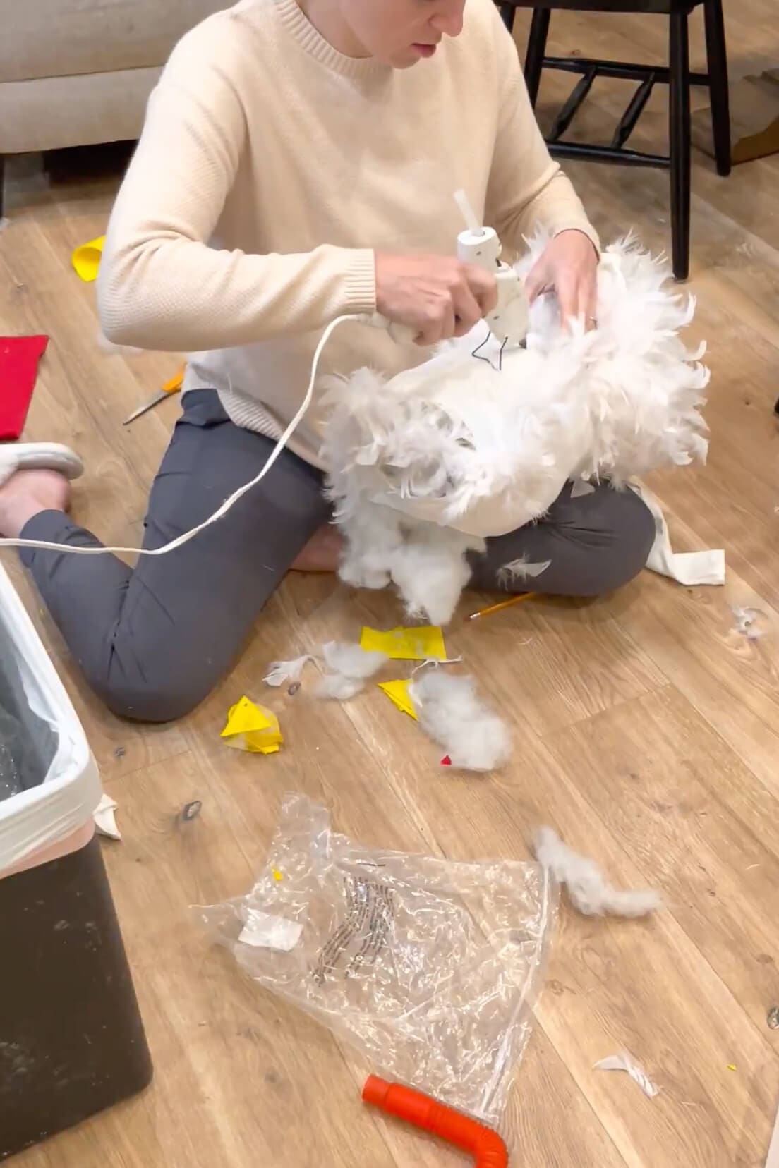 Mom using a hot glue gun to attach white feathers to the body of a handmade chicken costume.