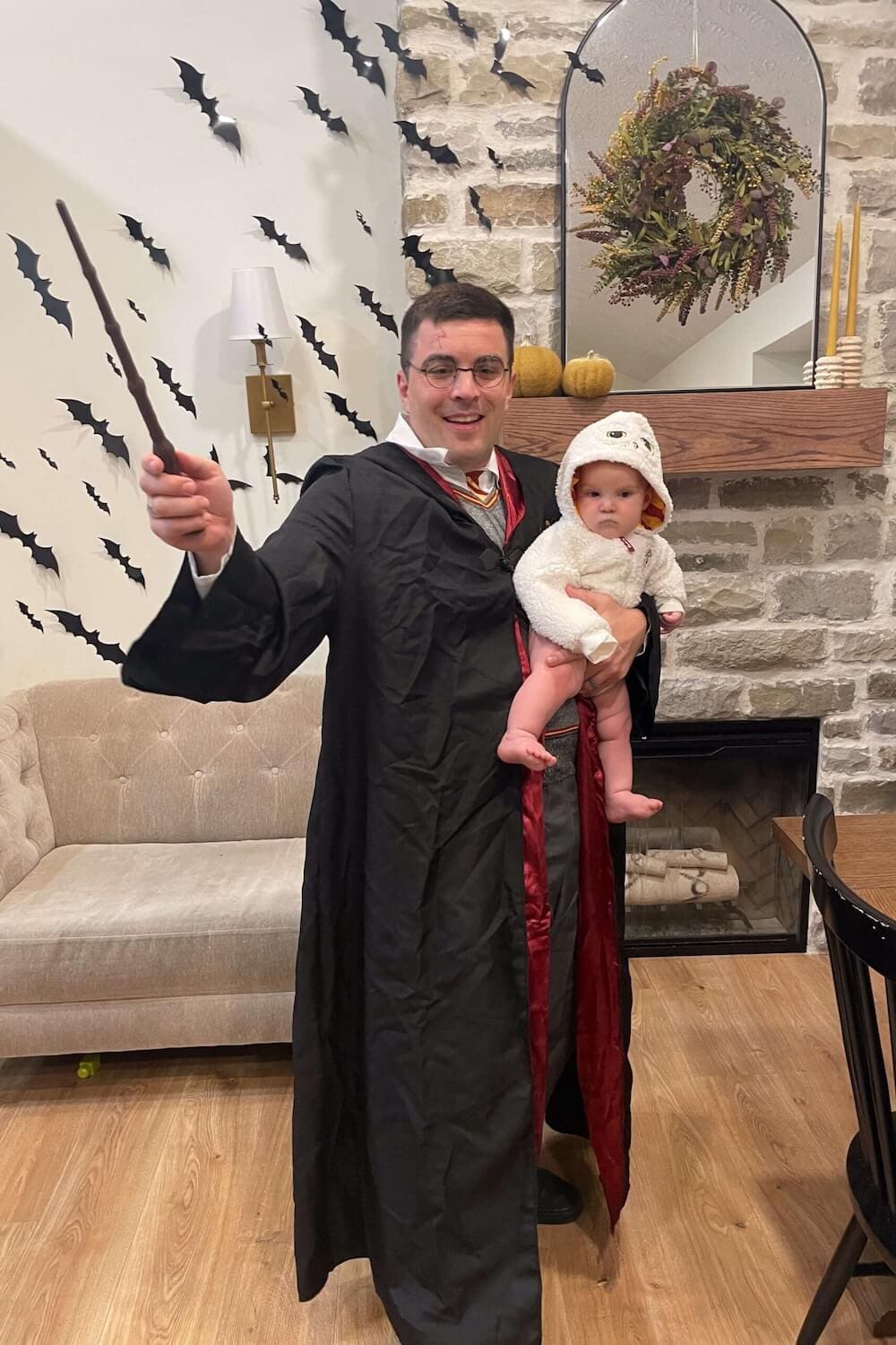 Father in Harry Potter costume with wand raised, holding baby dressed as Hedwig owl for Halloween.