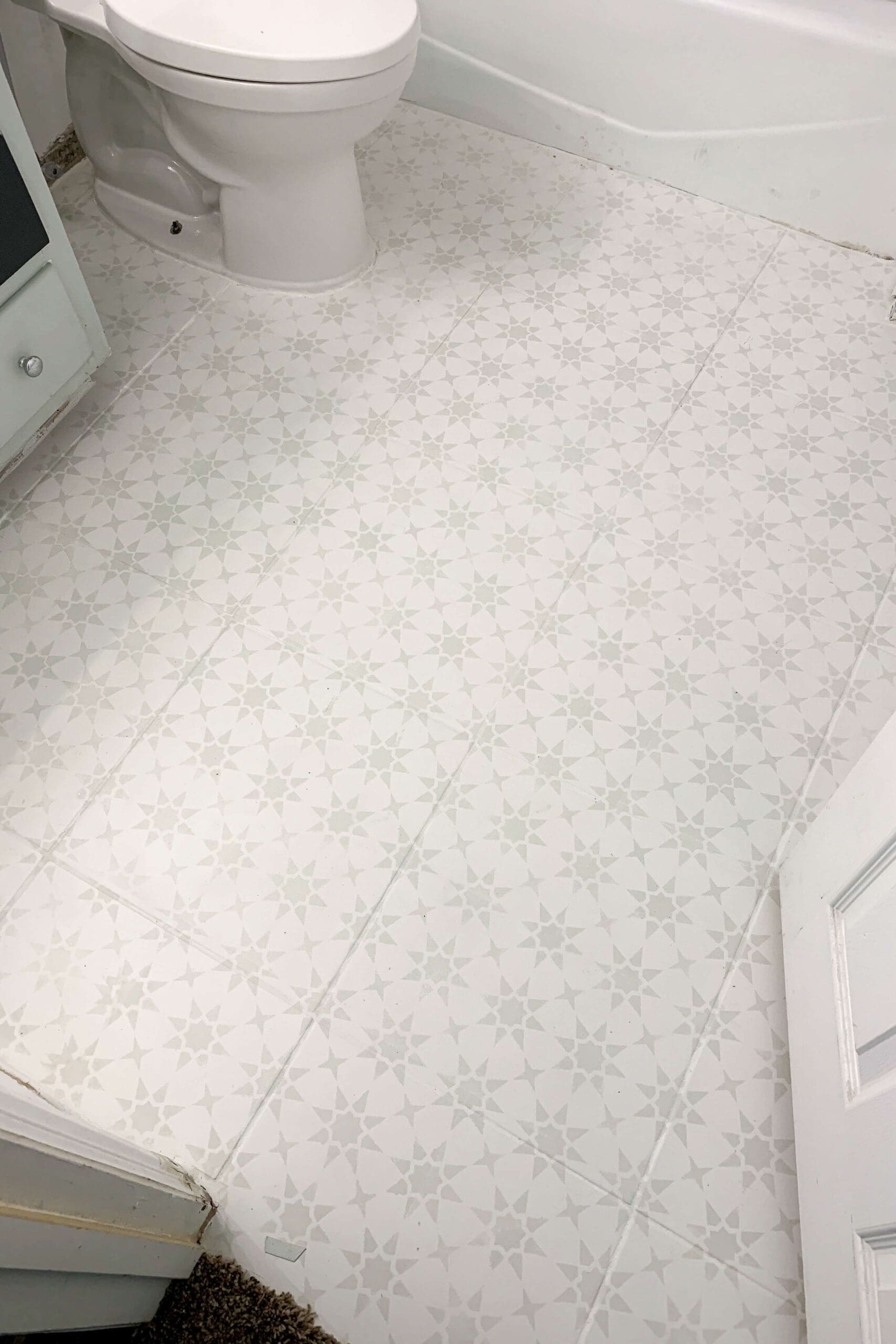 Full view of a small bathroom floor makeover showing freshly painted tiles with a repeating star stencil design, extending from the doorway around the toilet and bathtub.