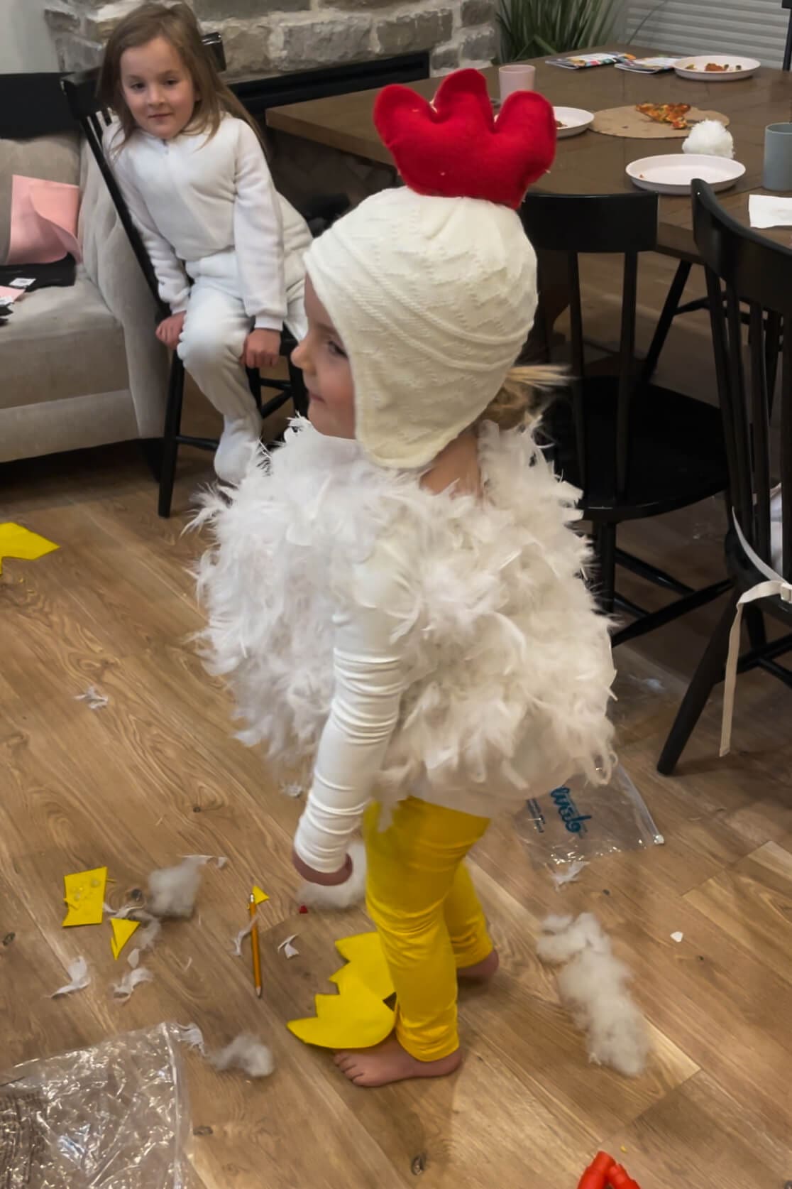 Side view of a child wearing a handmade chicken costume with white feathers, yellow leggings, and a knit hat with a red felt comb.