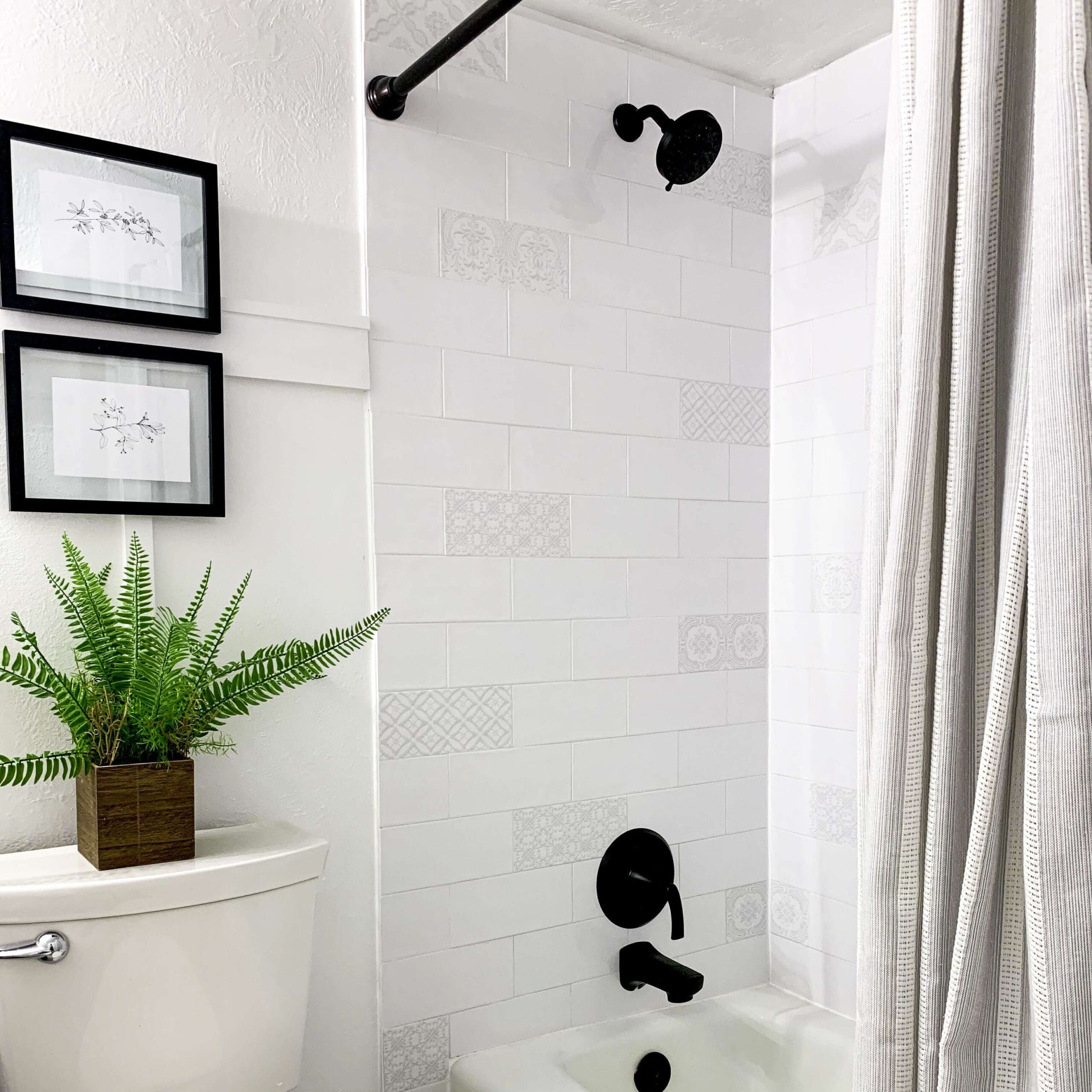 Freshly tiled shower wall with white subway tiles and soft patterned accent tiles, matte black shower fixtures, and framed botanical art above a toilet with a potted fern.
