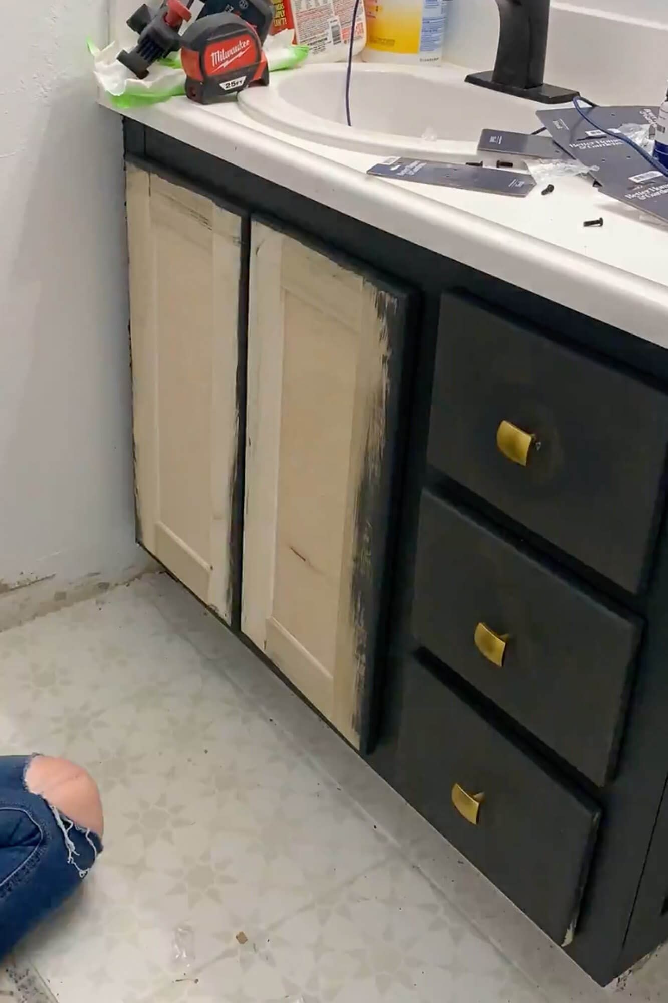 Bathroom vanity mid-makeover with new shaker-style doors in primer and dark painted drawers with gold hardware.