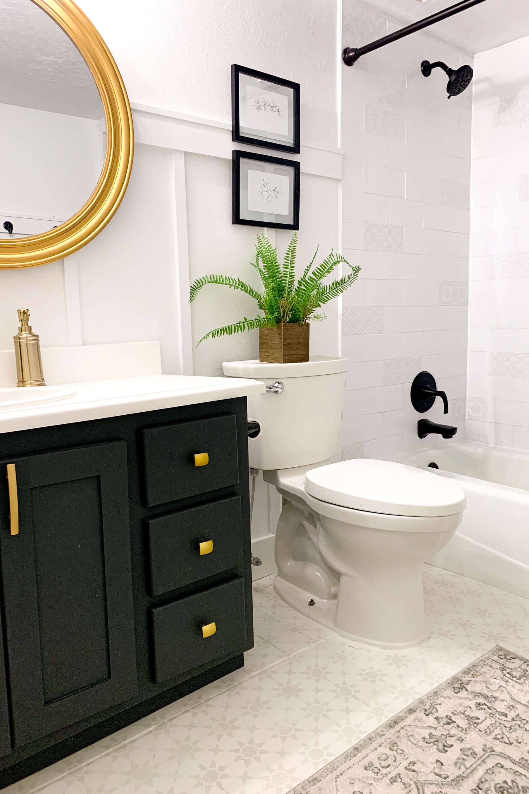 Cozy bathroom corner with stenciled tile floor, black vanity with gold hardware, potted fern, round gold mirror, and black shower fixtures.